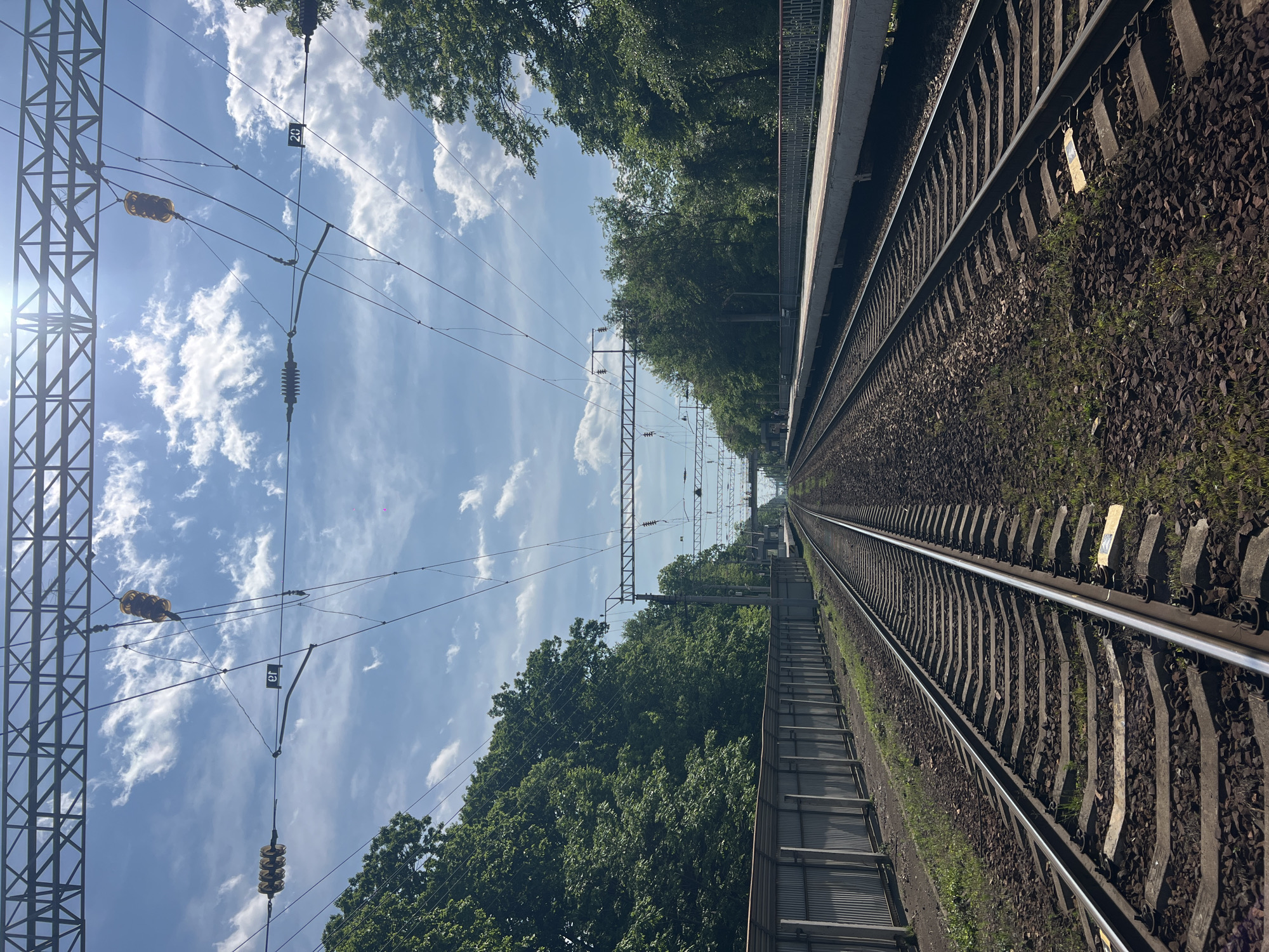 The image shows a railway track in Vladivostok, Russia. The tracks extend into the distance, flanked by dense green trees on both sides. Overhead power lines and support structures are visible against a blue sky dotted with scattered white clouds. A railway platform is seen on one side of the tracks.