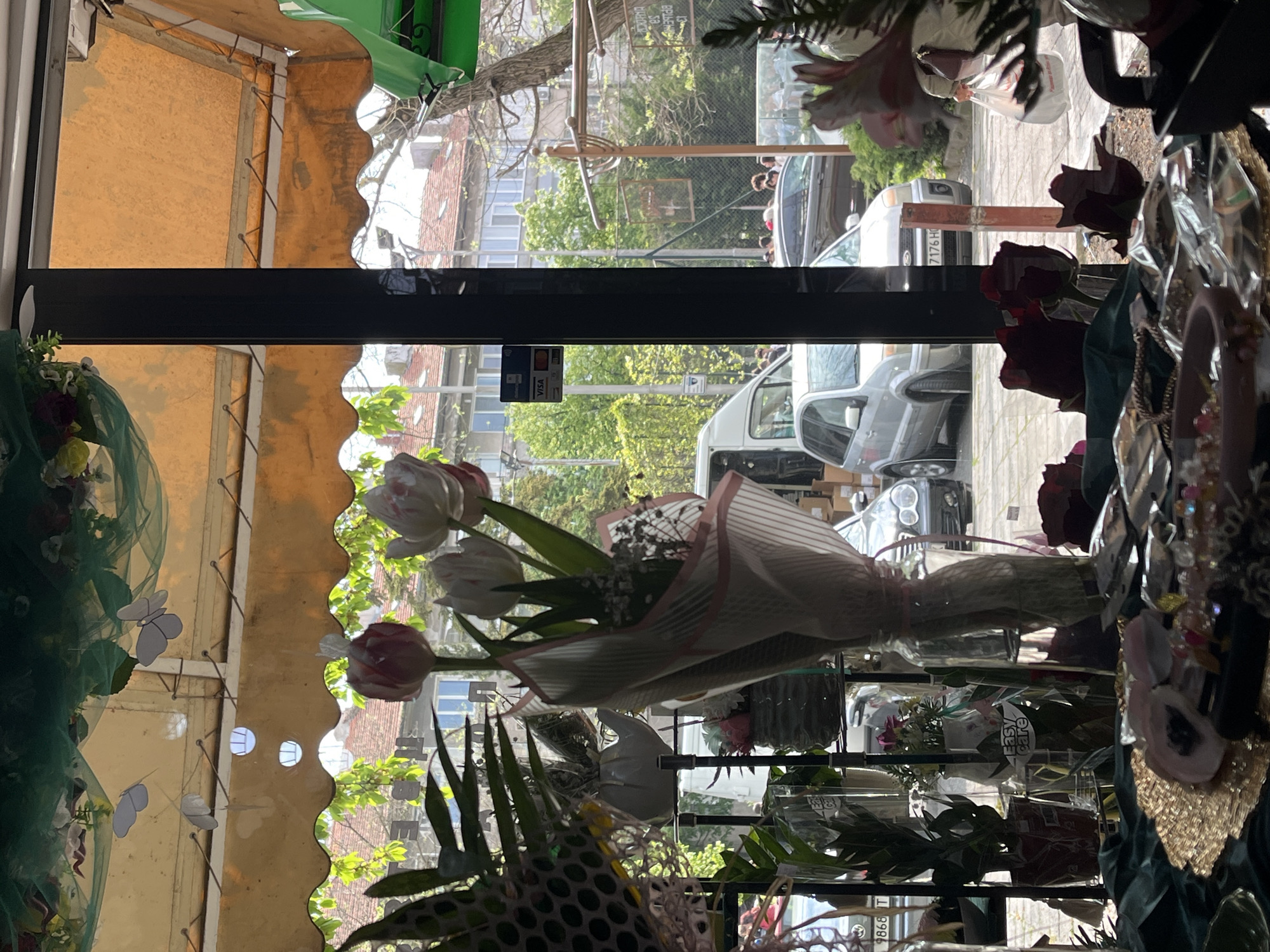 A flower shop display in Burgas, Bulgaria. The view is from inside the shop looking out through a window. A bouquet of tulips wrapped in paper sits on a shelf. Outside the window, a street with cars and pedestrians is visible. An awning is over the window. A credit card sign is visible on the inside of the window.