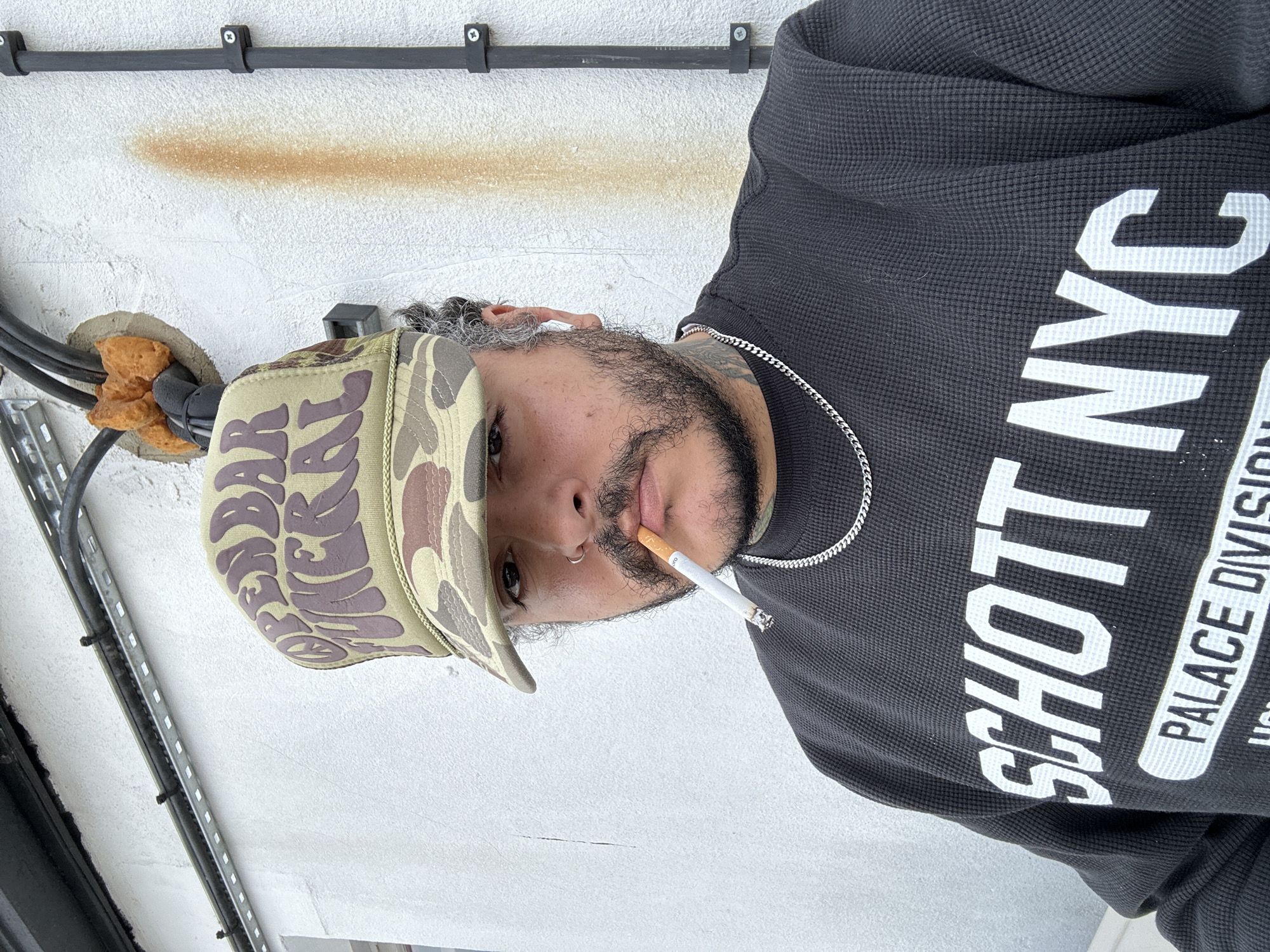 A man is facing the camera while holding a cigarette in his mouth. He is wearing a black waffle knit shirt that says "SCHOTT NYC PALACE DIVISION" in white capital letters. The man is also wearing a camouflage hat with lettering and a silver chain necklace. Cables run along the wall behind the man. There is a brown stain on the white wall. The location appears to be outdoors.