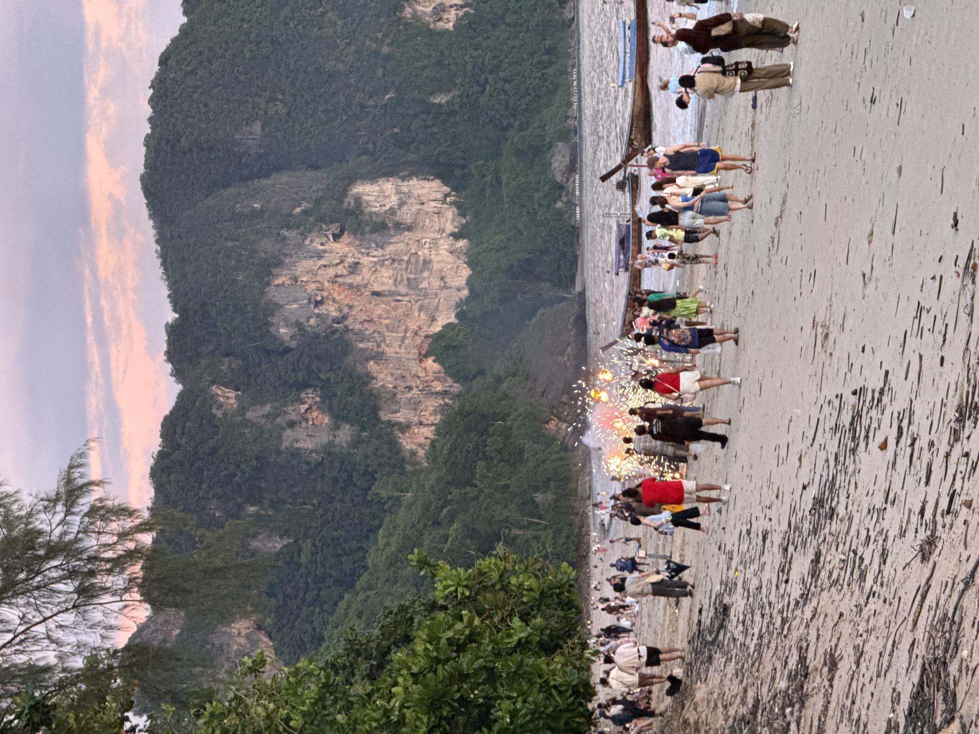 The image depicts a beach scene in Ban Klong Haeng, Thailand, during sunset. A crowd of people is gathered on the sand, watching fireworks ignite over the water. The beach is strewn with driftwood and debris. In the background, lush green mountains rise from the sea, with a pastel-colored sky above. Several longtail boats are moored offshore.