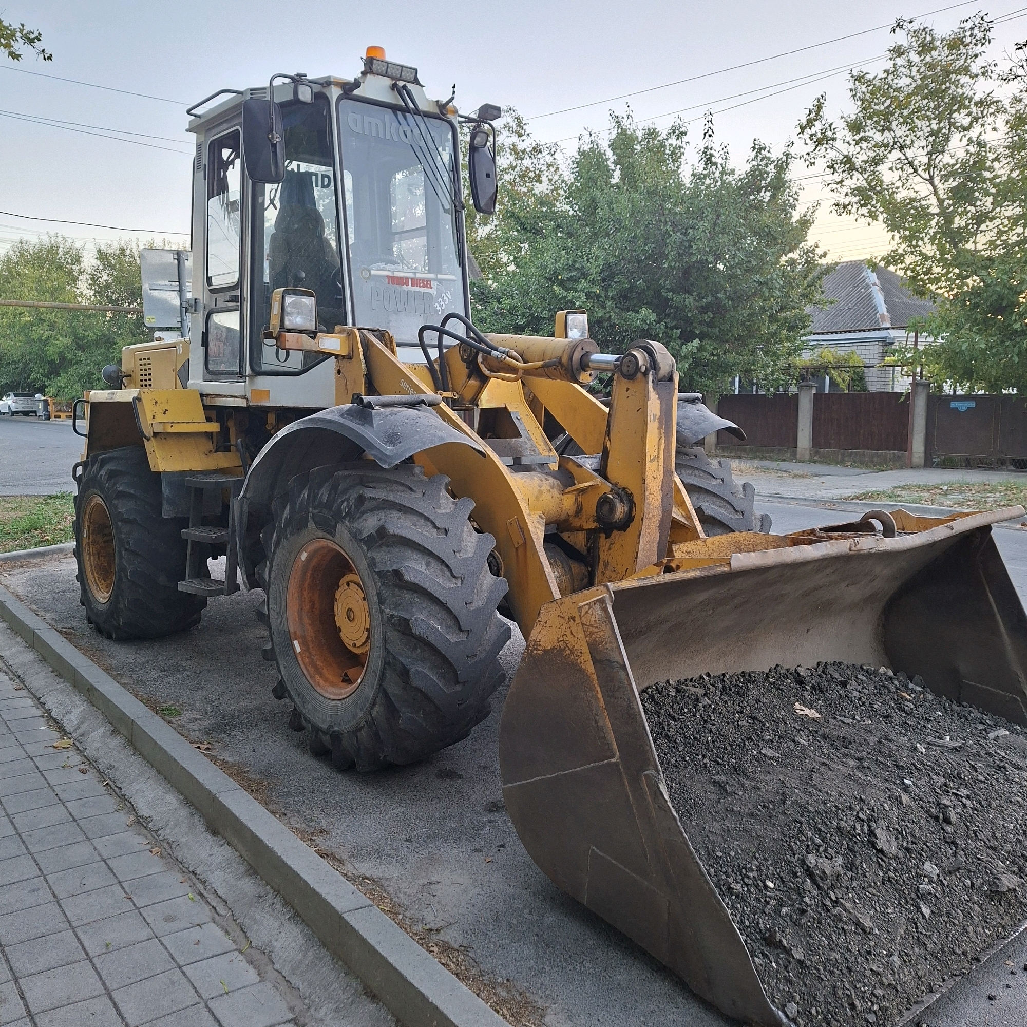 The image shows a yellow and white wheel loader parked on a paved surface next to a sidewalk with gray square tiles. The loader's large bucket is filled with dark material, likely asphalt or soil. The cab of the loader is visible, with windows reflecting the surroundings. Trees and a house with a gray tiled roof are in the background. The sky is overcast, suggesting it is either early morning or late afternoon. There are power lines visible in the background, and a street with a white car parked on the left side of the image. The scene appears to be in a residential or semi-urban area. The loader is positioned on the right side of the frame, with its front facing towards the viewer.