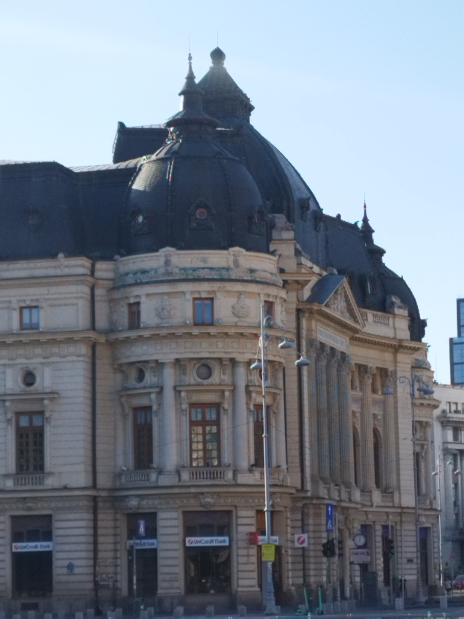 The image shows the UniCredit Bank building in Bucharest, Romania. The building is a large, ornate structure with a dark, domed roof and classical architectural elements. The facade is made of light-colored stone, and there are numerous windows and decorative carvings. In the foreground, there is a street with traffic lights and street lamps. A few people can be seen walking on the sidewalk. The sky is clear and blue.