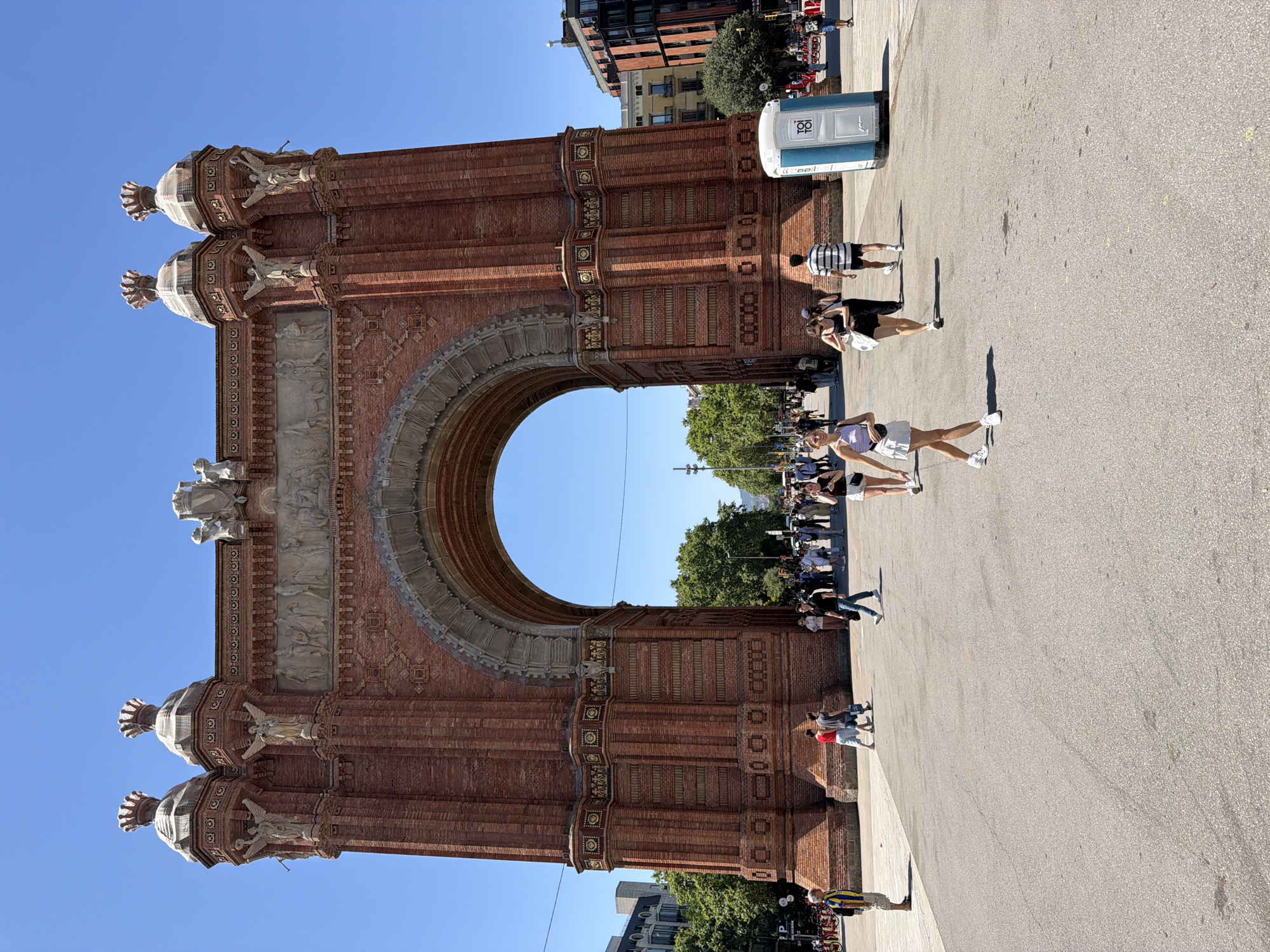 The image shows the Arc de Triomf in Barcelona, Spain, on a sunny day. The grand brick archway dominates the center of the frame, with intricate carvings and decorative elements visible. Several people are walking in the plaza in front of the arch. In the foreground, a woman in a white skirt and lavender top walks towards the camera. To her left, a woman in a black tank top and shorts walks away from the camera. Further in the background, other individuals and small groups can be seen milling about. A blue portable toilet is positioned to the right of the arch. Trees line the sides of the plaza, and buildings are visible in the distance. The sky is clear and blue.