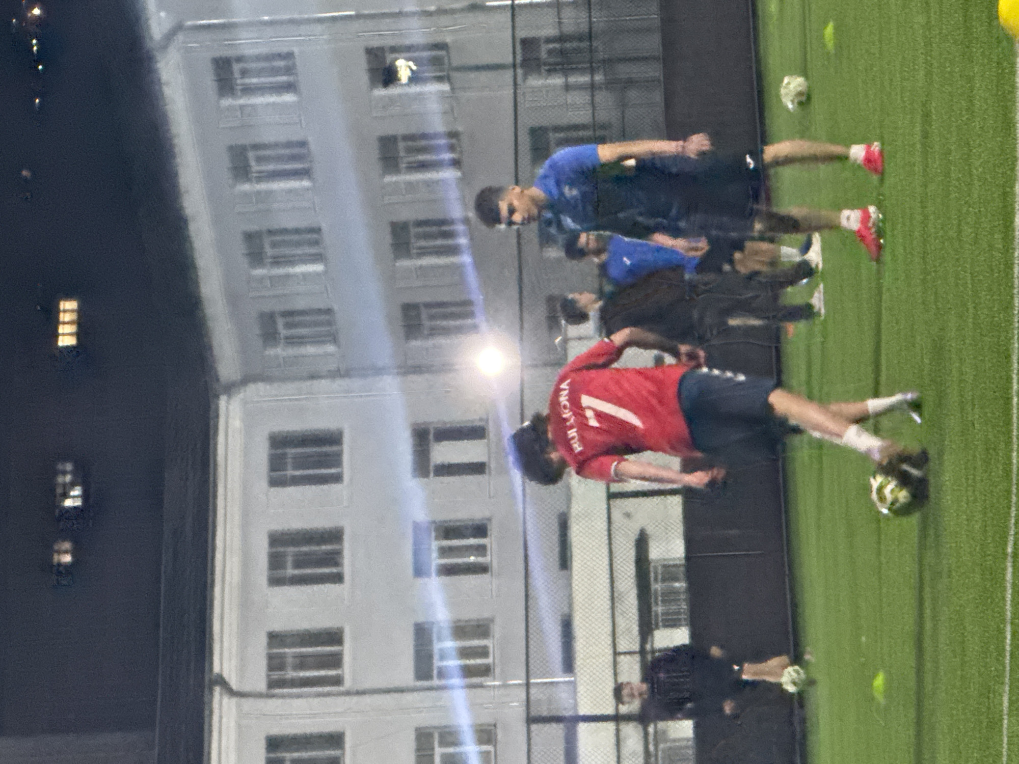 The image shows a group of people playing soccer on a green field at night, with an adjacent building complex suggesting an urban setting in Tbilisi, Georgia. At least two teams are present, identifiable by their differently colored uniforms: one in red and one in blue. One player in red is closer to the camera, with a soccer ball at their feet. Another is in blue. Additional people are scattered around the soccer field, seemingly engaged in the same activity. The soccer field is surrounded by a fence, and nearby a multi-story building can be seen with lit windows.