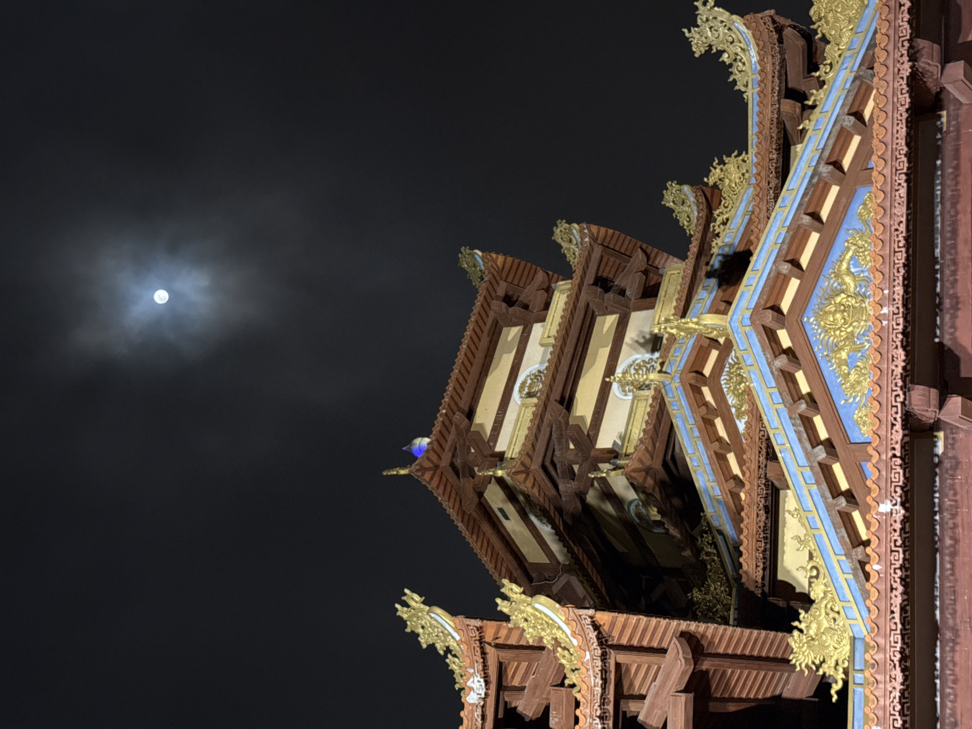 The image depicts a section of the Linh Ứng Pagoda in Đà Nẵng, Vietnam, at night with a visible full moon. The pagoda’s ornate roof structure dominates the right side of the frame, showcasing intricate carvings, gold embellishments, and detailed paintwork in shades of blue, brown, and gold. The night sky is dark, with the moon visible on the left side of the frame, partially obscured by clouds.