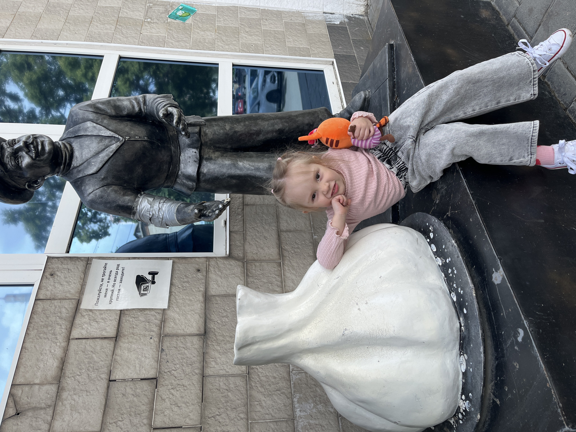A young child is seated outdoors in front of a dark-colored statue and a large white garlic sculpture, against the backdrop of a building with tiled walls and reflective windows, in the city of Unknown, Unknown.
The child, appearing to be a toddler or preschooler, has light brown hair tied in two small pigtails with pink hair ties. She is wearing a light pink long-sleeved ribbed top, faded grey wide-leg jeans with a ruched elastic waistband, white low-top sneakers with red trim, and pink socks. She holds an orange and striped plush toy in her left hand and rests her right arm on the white garlic sculpture. She is smiling and looking directly at the camera, with her left hand clenched near her chin.
To the child's right, a dark-colored statue of a man stands. The man wears a cap, a suit jacket over a shirt and tie, and has a prominent mustache. His proper left arm (viewer's right) is wrapped with silver duct tape, while his proper right hand is extended forward with an open palm. The statue appears to be gazing upwards and slightly to its right.
To the child's left, a large, white sculpture shaped like a bulb of garlic sits on a round, black base. The garlic sculpture shows signs of weathering with visible cracks and paint loss. The black base is splattered with white marks. Both the statue and the garlic sculpture are situated on a dark, elevated platform.
Behind the child and sculptures, the building's exterior features light grey rectangular tiled walls. Large, blue-tinted reflective windows are set into the wall, showing reflections of green trees and a white sedan. A rectangular white sign, affixed to the tiled wall above the garlic sculpture, displays Russian text and a black icon of a CCTV camera. A smaller, partially obscured green and white sticker, possibly a UPS logo, is visible on the wall to the right of the statue. The ground directly in front of the platform is paved with dark material.