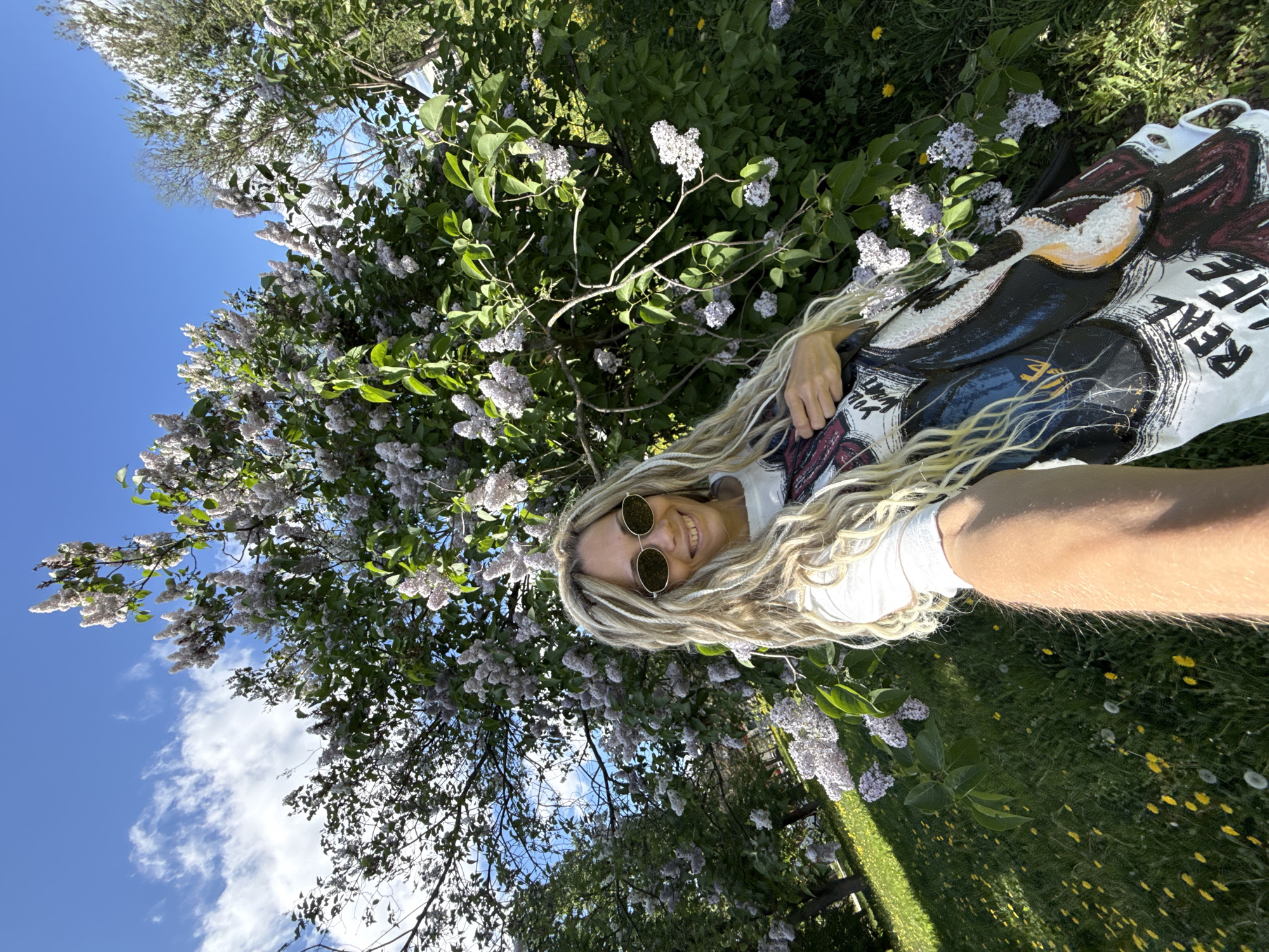 A woman with blonde dreadlocks and round sunglasses is lying in the grass, seemingly taking a selfie. She is wearing a white T-shirt with a graphic design. The location appears to be outdoors, possibly a garden or park in Шебунино, Russia. There are lilac bushes with white and purple flowers surrounding her, and the sky is visible with a few clouds. The grass is green, and there are dandelions scattered around.