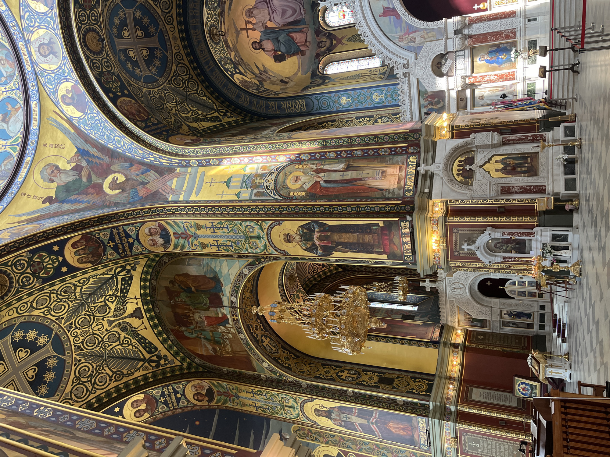 The image shows the interior of the Ascension Cathedral in Novocherkassk, Russia. The cathedral features ornate gold and blue decorations on its vaulted ceilings, adorned with religious frescoes and mosaics. A large, multi-tiered chandelier hangs in the center, illuminated by numerous candles. The walls are decorated with religious icons and paintings, depicting biblical scenes and figures. In the foreground, the checkered floor leads towards an altar area with an ornate iconostasis. Red velvet ropes separate the main floor from the altar. The overall impression is one of grandeur and religious devotion.