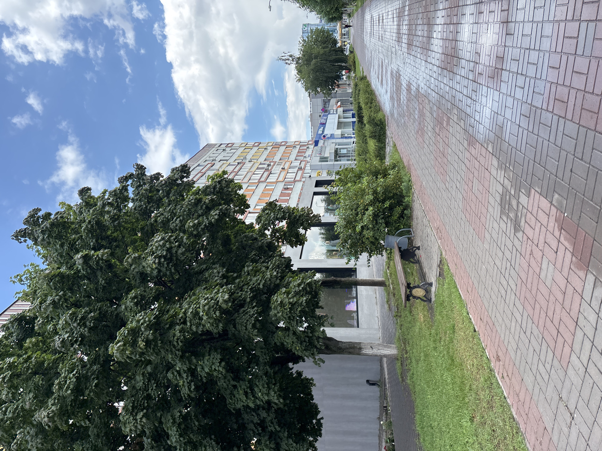 The image captures a scene on a pedestrian walkway in Naberezhnye Chelny, Russia. The walkway is paved with red and grey bricks, with some areas appearing wet, suggesting recent rain. To the left of the walkway, there is a patch of green grass and a large, leafy tree with a grey-painted trunk. Further left, a multi-story building with a gray facade is visible, featuring large windows and a few trees planted in front of it. To the right of the walkway, a long, multi-story residential building with a colorful facade of orange, yellow, and white sections dominates the background. Several shops with large glass windows are located on the ground floor of this building, with various signs displaying company names. In the foreground of the walkway, a wooden bench with ornate metal legs is placed on the grass. Further down the walkway, a few individuals can be seen walking. The sky above is a mix of blue with scattered white clouds.
