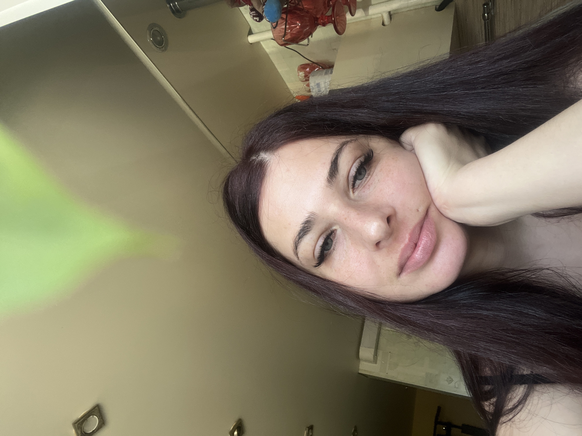 A woman with dark hair and light skin is posing for a selfie. She is resting her head on her hand. She appears to be indoors, possibly in a bathroom or bedroom, based on the background, which includes a light-colored wall, some fixtures, and what appear to be decorative items on a shelf. The specific location in Chaikovsky, Russia cannot be determined.