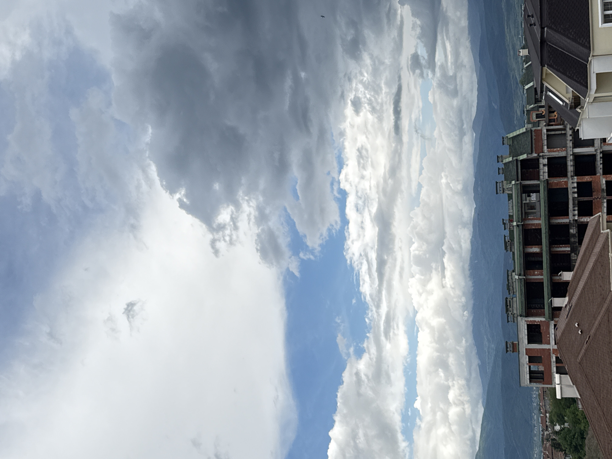 The image shows a partially constructed building in Plovdiv, Bulgaria, with a backdrop of mountains. The sky is filled with a mix of gray and white clouds, with a patch of blue visible. An open-frame building stands partially complete, with brickwork visible. Adjacent to the building, the rooftops of existing structures are shown.