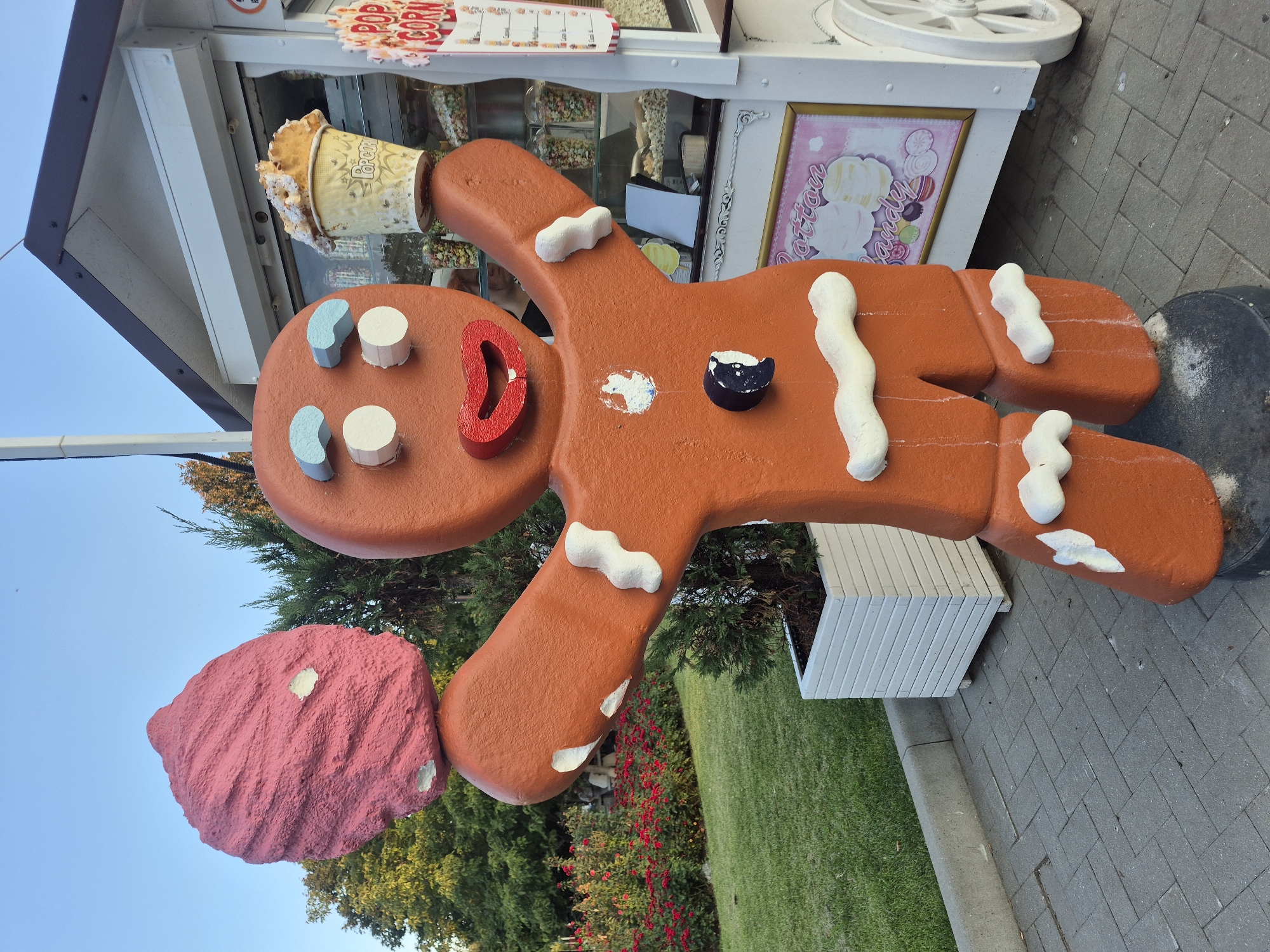 The image depicts a large, bright orange, gingerbread man statue standing on a paved walkway in front of a white food cart. The statue has blue icing eyebrows, white circular eyes, and a red smiling mouth. It is holding a large pink, cotton candy-like object in its left hand and a cup of popcorn, or a similar snack, in its right hand. The food cart behind the statue is selling popcorn and other sweets, with a "POP CORN" sign visible. The environment appears to be a park or outdoor recreational area, with a lush green lawn and flowering bushes in the background. The sky is clear and blue. The overall scene suggests a cheerful, festive atmosphere, possibly in an amusement park or a similar public space. Based on available information, this scene is located in Chișinău, Moldova.