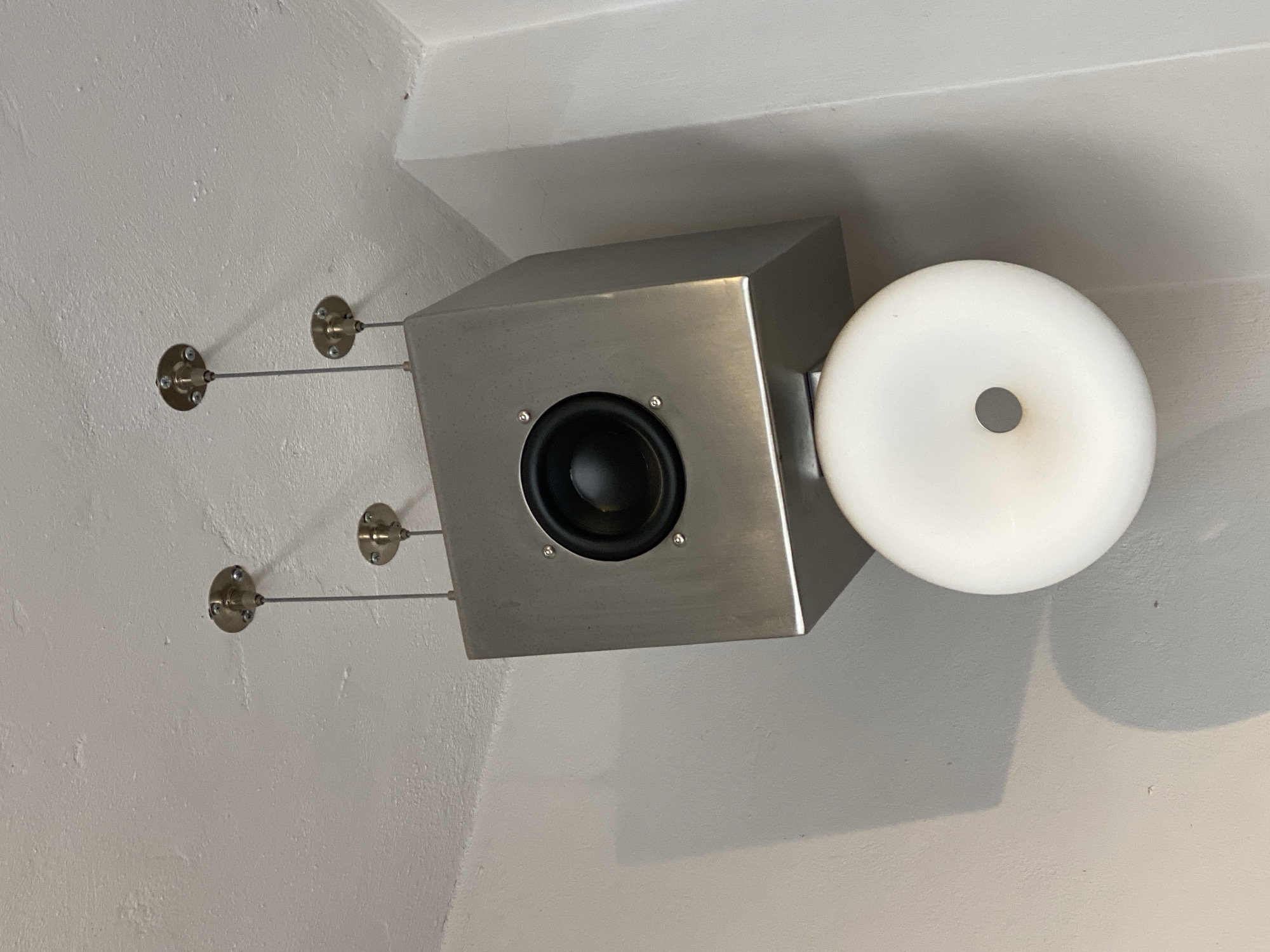 The image shows a silver, cube-shaped speaker mounted on a white wall. The speaker is attached to the wall using two sets of cables connected to circular mounts. A white, donut-shaped object is attached to the side of the speaker. The environment appears to be an interior space with white walls and a ceiling. The image was taken in Puebla, Mexico.