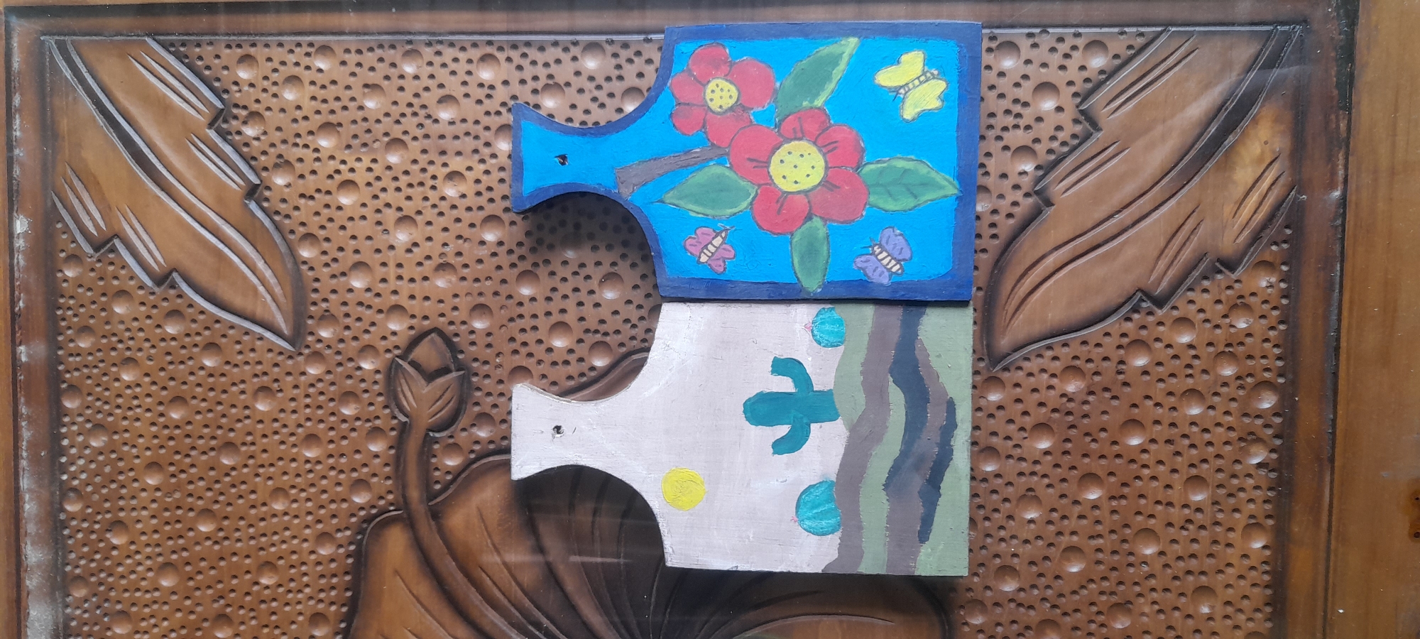 Two decorative wooden items, possibly small cutting boards or wall hangings, are placed side-by-side on a textured wooden surface. The item on the left is painted with a desert scene featuring a yellow sun, a green cactus, and blue rounded hills against a light tan sky. The item on the right is painted with a vibrant blue background depicting red flowers with yellow centers and green leaves, as well as three butterflies in pink and yellow. The wooden background has a carved pattern of small raised circles and stylized leaf motifs. The image is taken from a slightly overhead perspective, and the overall lighting is neutral, suggesting an indoor setting without direct sunlight. There are no people or animals visible in the image, and no specific geographical markers are present to indicate a location in Iser, Indonesia or elsewhere.