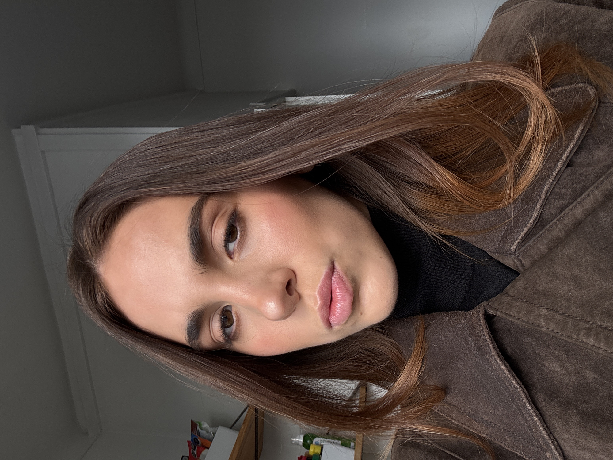 The image displays a headshot-style portrait of a female individual with long, straight dark brown hair featuring auburn highlights, parted slightly off-center. She has fair skin, prominent dark eyebrows, brown eyes, and her lips are naturally pink and slightly parted. She is wearing a dark brown collared jacket, appearing to be suede or a similar material, layered over a black ribbed turtleneck sweater. The individual is positioned centrally, looking directly into the camera with a neutral expression.
The background is an indoor setting. To the left, a light-colored wall features wooden shelving holding miscellaneous items, including a white rectangular container with several small packets and a green spray bottle. A darker gray wall section is visible on the right side of the frame. White molding is present near the ceiling on the upper left. No identifiable landmarks, specific architectural features, or unique environmental cues are present to determine the precise geographical location or to confirm it is within the city of Paris, France.