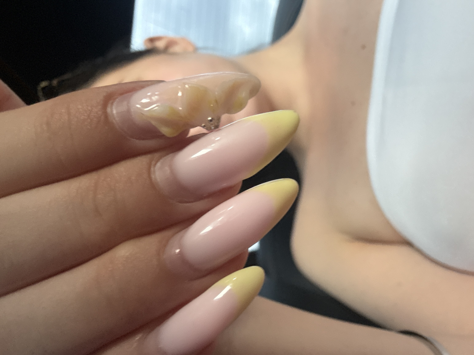 The image is a close-up, high-angle shot of a person's hands and nails, with the background blurred. The nails are long and almond-shaped, with a light pink base and a yellow French tip. One nail has an abstract design with yellow and clear acrylic. The person appears to be wearing a white strapless top. The background suggests an indoor setting with soft lighting, possibly a car interior or a room. There is no indication of the location being Burgas, Bulgaria.
