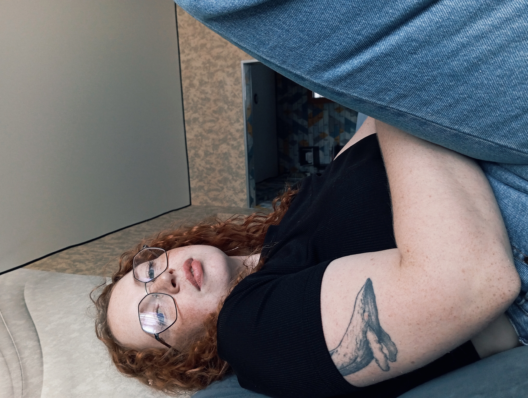 An upward-angled shot captures a person with fair skin, numerous freckles, and curly reddish-brown hair, lying down in an indoor setting. They are wearing a black ribbed short-sleeved top and hexagonal-framed glasses, through which their blue eyes are visible. Their lips are slightly parted. A prominent tattoo of a whale or similar marine creature is visible on their right upper arm.
The person appears to be resting on a light-colored, upholstered surface, possibly a couch or bed. Partially visible in the foreground are blue denim jeans, likely belonging to the subject.
The background shows a light grey wall or screen on the left. To the right, a wall with a mottled, textured wallpaper in shades of brown and grey is visible. Through an opening, possibly a doorway, into another room, distinctive patterned tiles or wallpaper with blue, orange, and white geometric designs can be seen. The lighting in the image is soft and even, suggesting a daytime interior. There is no visible text in the image. The scene depicts a casual, relaxed moment within what appears to be a home in Yurgamysh, Russia.