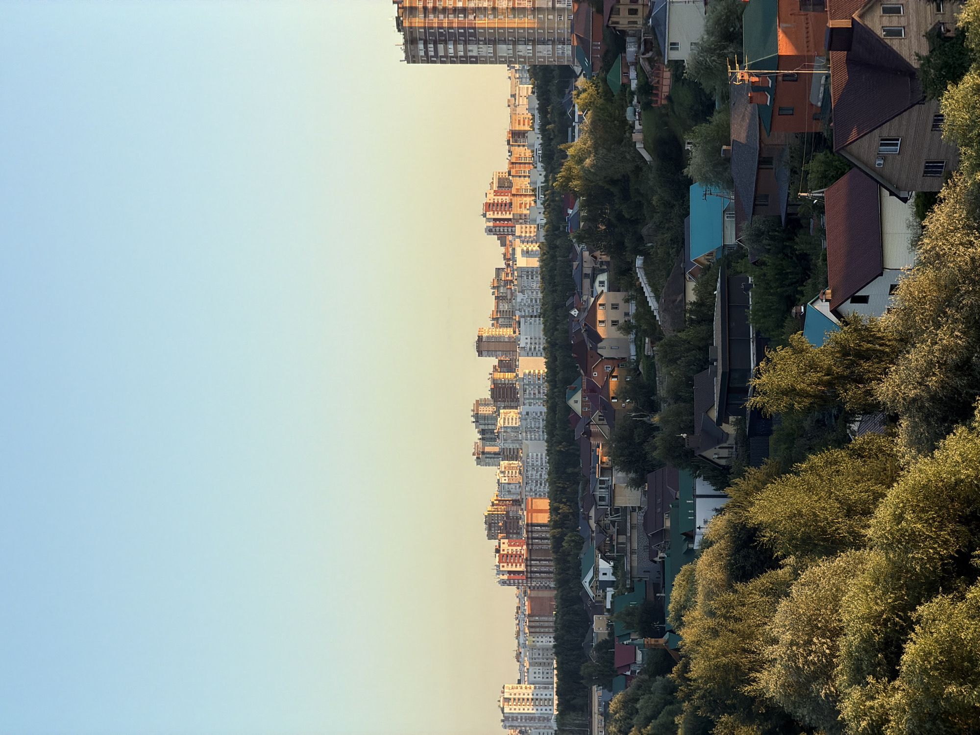 An aerial, high-angle, full shot captures a city skyline and a suburban area under a clear, pale blue sky with a hint of yellow light, suggesting early morning or late afternoon. In the distance, a dense cluster of modern, multi-story buildings forms the city skyline. Closer to the foreground, a sprawling suburban landscape unfolds, characterized by numerous detached houses with pitched roofs, varying in color and architectural style. These homes are nestled amongst lush green trees and foliage, creating a sense of a community integrated with nature. A tall, narrow apartment building, with visible windows, stands prominently on the right side of the frame, adding a vertical element to the suburban scene. The overall impression is a transition from urban density to a more relaxed, residential setting.