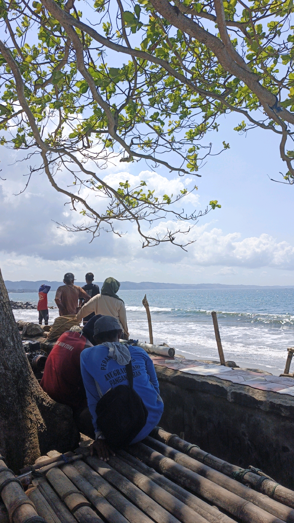 This image depicts a group of individuals on a beach in Desa Pagandaran, Indonesia. The scene is dominated by the ocean, with gentle waves rolling onto the shore under a bright, cloudy sky. Lush green tree branches with sparse leaves frame the upper portion of the image, suggesting a natural, coastal environment.
In the foreground, several people are seated on a bamboo structure, facing away from the camera and towards the sea. One individual wears a blue long-sleeved shirt with text printed on the back, and a black bag is slung over their shoulder. To their left, another person is wearing a red garment.
Further back, near the water's edge, more individuals are visible. One person in a red shirt and dark pants stands looking out at the sea. Two other individuals are also standing, one wearing a brown shirt and the other a black shirt. A person in a beige top and headscarf is also present, facing the ocean.
The shoreline is rocky in some areas, with the ocean meeting a paved or tiled pathway in others. There are wooden poles and some rope or fishing equipment scattered near the water. The overall atmosphere suggests a casual gathering or resting spot for locals by the sea.