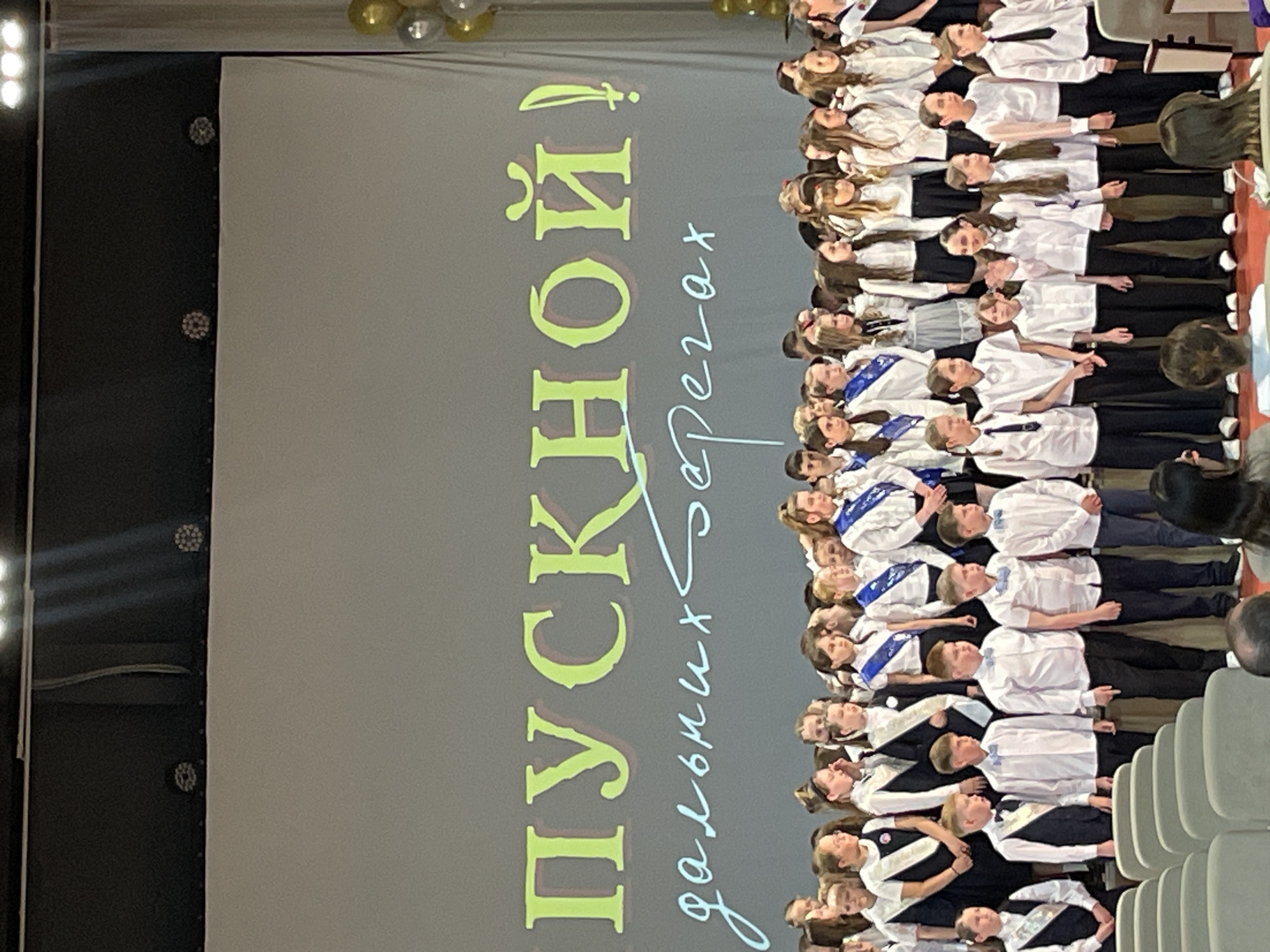 The image shows a group of children lined up side-by-side in rows. They are wearing white shirts and dark pants, and some have sashes or ribbons. A screen behind them has text in Russian that includes the word "Пушкин" (Pushkin). The background and seating suggest a stage or auditorium setting. The location is implied to be in the city of Izhevsk, Russia. The children appear to be participating in a performance or event.
