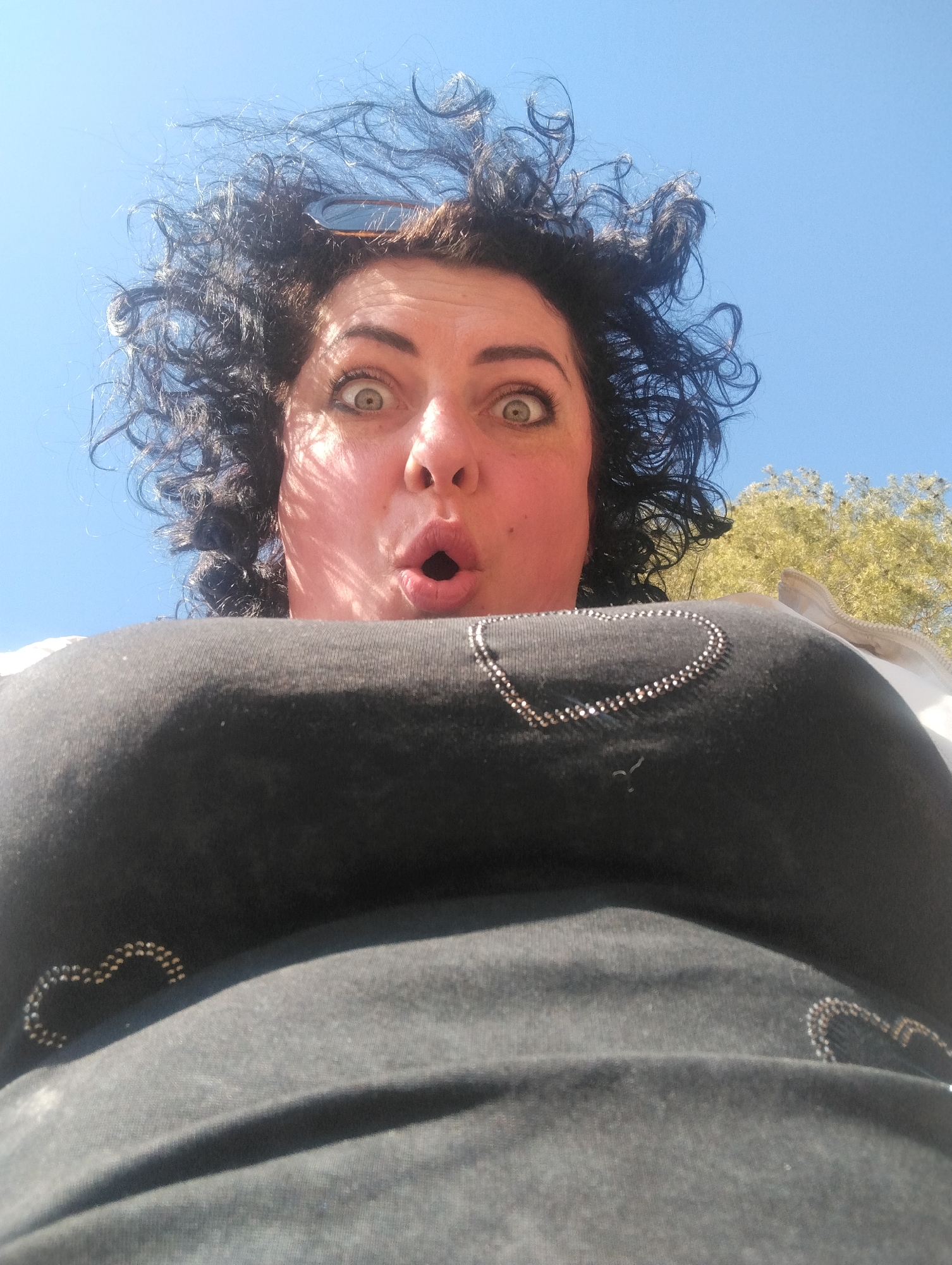 A woman with dark, curly hair is photographed from a low angle against a clear blue sky and partial tree foliage. She is wearing a black top with a heart-shaped rhinestone embellishment on the chest. Her mouth is open in a surprised expression and her eyes are wide. The photograph appears to have been taken outdoors during the day. There is no information available in the image to indicate this image was taken in the city of Votkinsk, Russia.