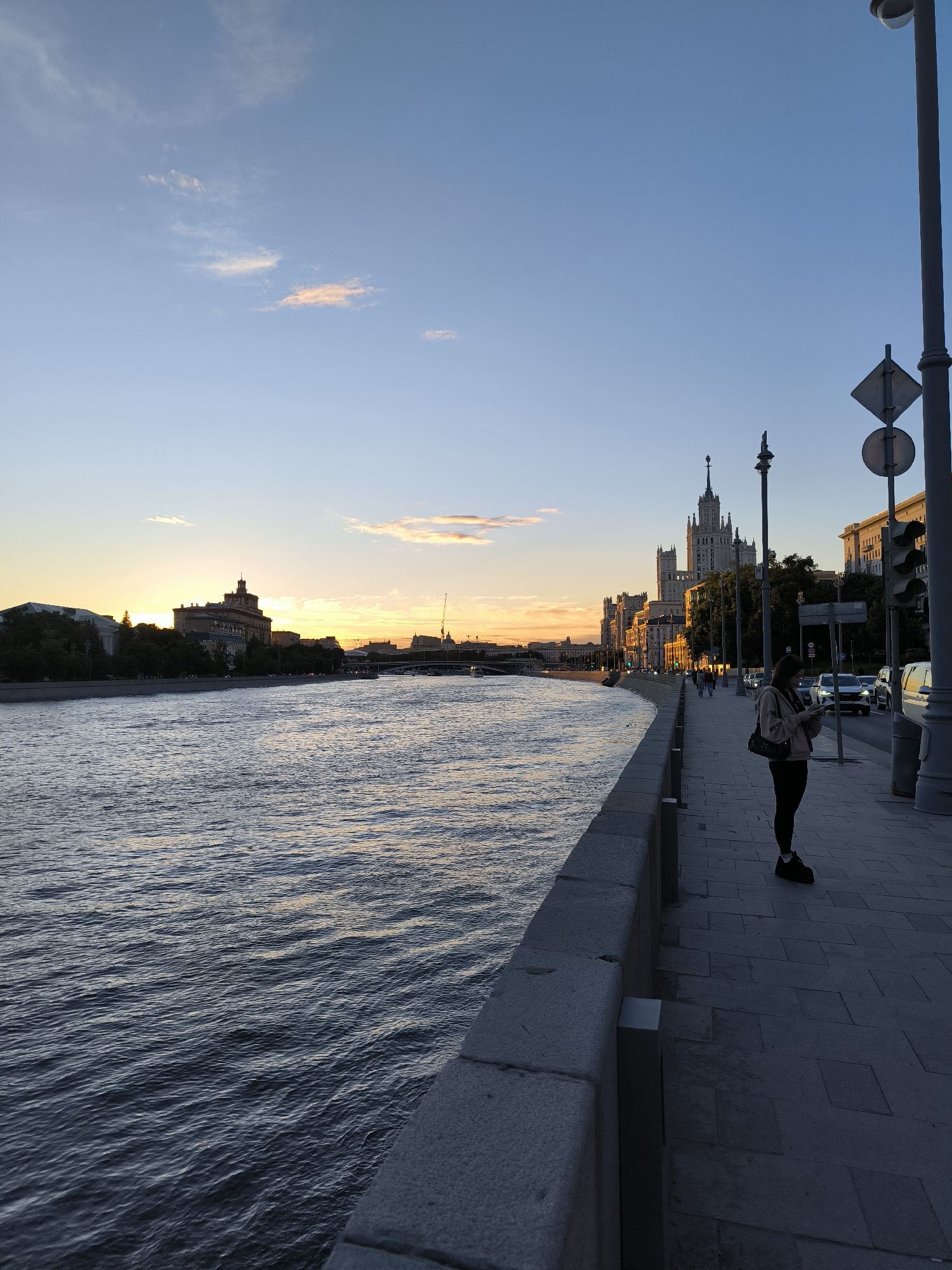 The image depicts a view of the Moskva River in Moscow, Russia, during sunset. On the left side of the frame, the river flows with gentle ripples, reflecting the warm colors of the sky. The opposite bank features a cityscape with buildings and trees silhouetted against the setting sun. On the right side of the frame, a woman stands on a paved pedestrian walkway beside the river. She is looking at her phone, wearing dark leggings and a light-colored top. Behind her, a tall, distinctive Stalinist-era skyscraper dominates the skyline, illuminated by the golden hour light. Several cars are visible on the road behind the walkway. Streetlights and traffic signals are also present. The sky is a gradient of blue and orange, with a few wispy clouds. The overall scene conveys a peaceful urban sunset atmosphere.