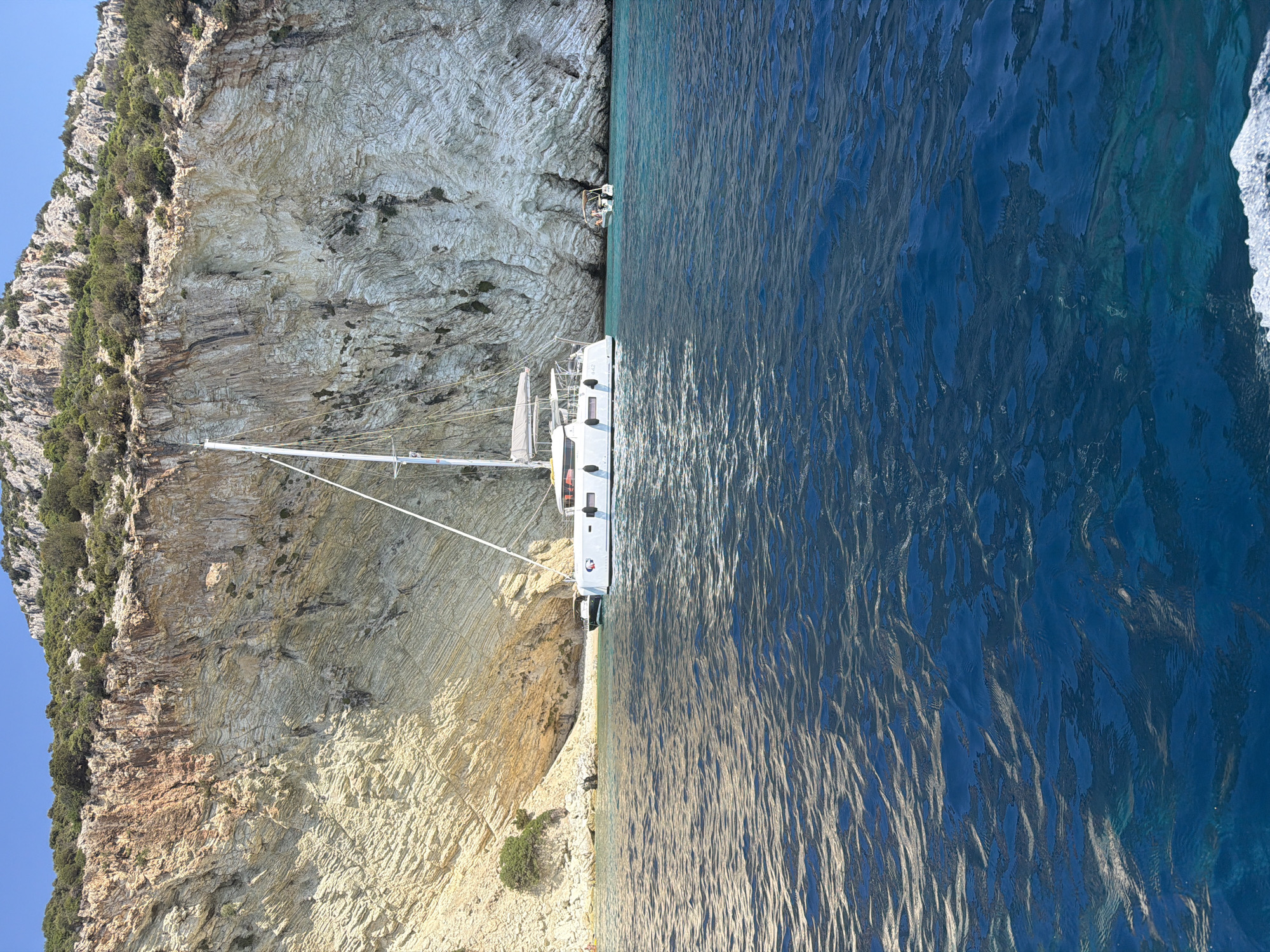 A catamaran is anchored in the clear blue waters of a cove, near a sheer, layered cliff face. The cliff is a mix of tan and grey rock, with sparse vegetation growing near the top. The water is calm and transparent, allowing a view of the seabed in shallower areas. A small dinghy is moored behind the catamaran. In the distance, at the base of the cliff, a small beach is visible with a few people on it. The sky is a clear, bright blue. The scene is located in the Municipal Unit of Kastos, Greece.