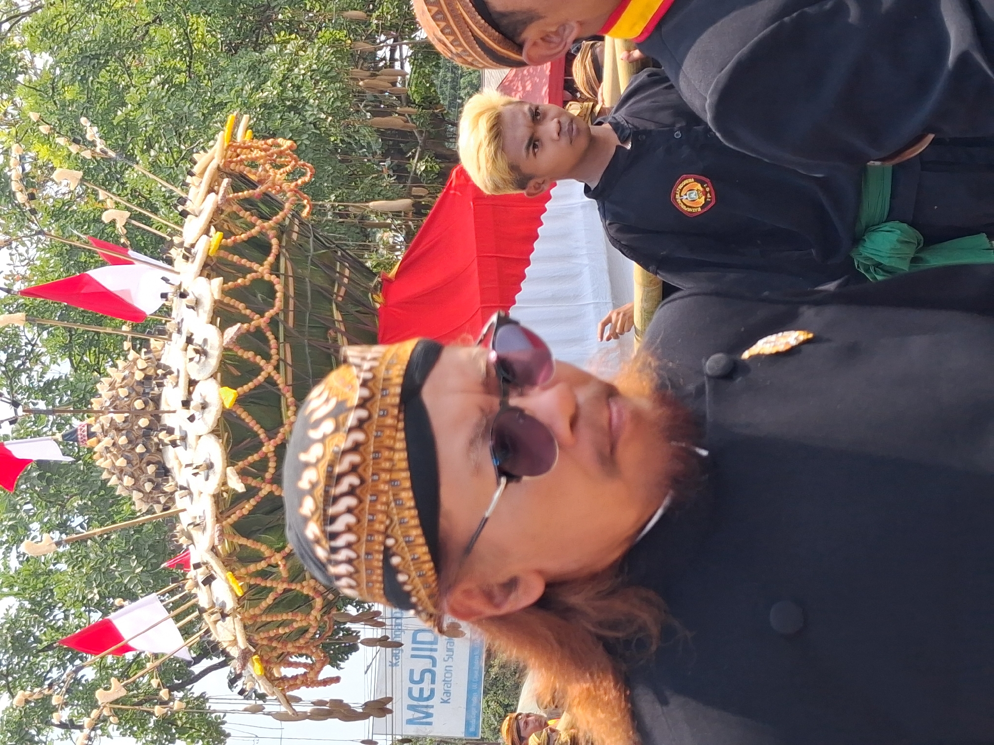 The image captures a street scene in Surakarta, Indonesia, likely during a parade or festival. In the foreground, a man with long, reddish-brown hair and a beard wears a black jacket, sunglasses, and a patterned Kufi. To his right, a young man with bleached blonde hair, also in a black uniform, stands behind a wooden railing. Further to the right, another individual in a similar black uniform with a yellow and red collar is partially visible. Behind the people, a large, elaborate structure adorned with red and white flags and what appear to be dried fruits or vegetables is prominently displayed. A red and white tent is also visible in the background, along with lush green trees and foliage. A sign that reads "MESJID" and "KARATON SURAKARTA" is partially visible to the left, indicating a location near the Surakarta Palace. The overall atmosphere suggests a cultural event or celebration.
