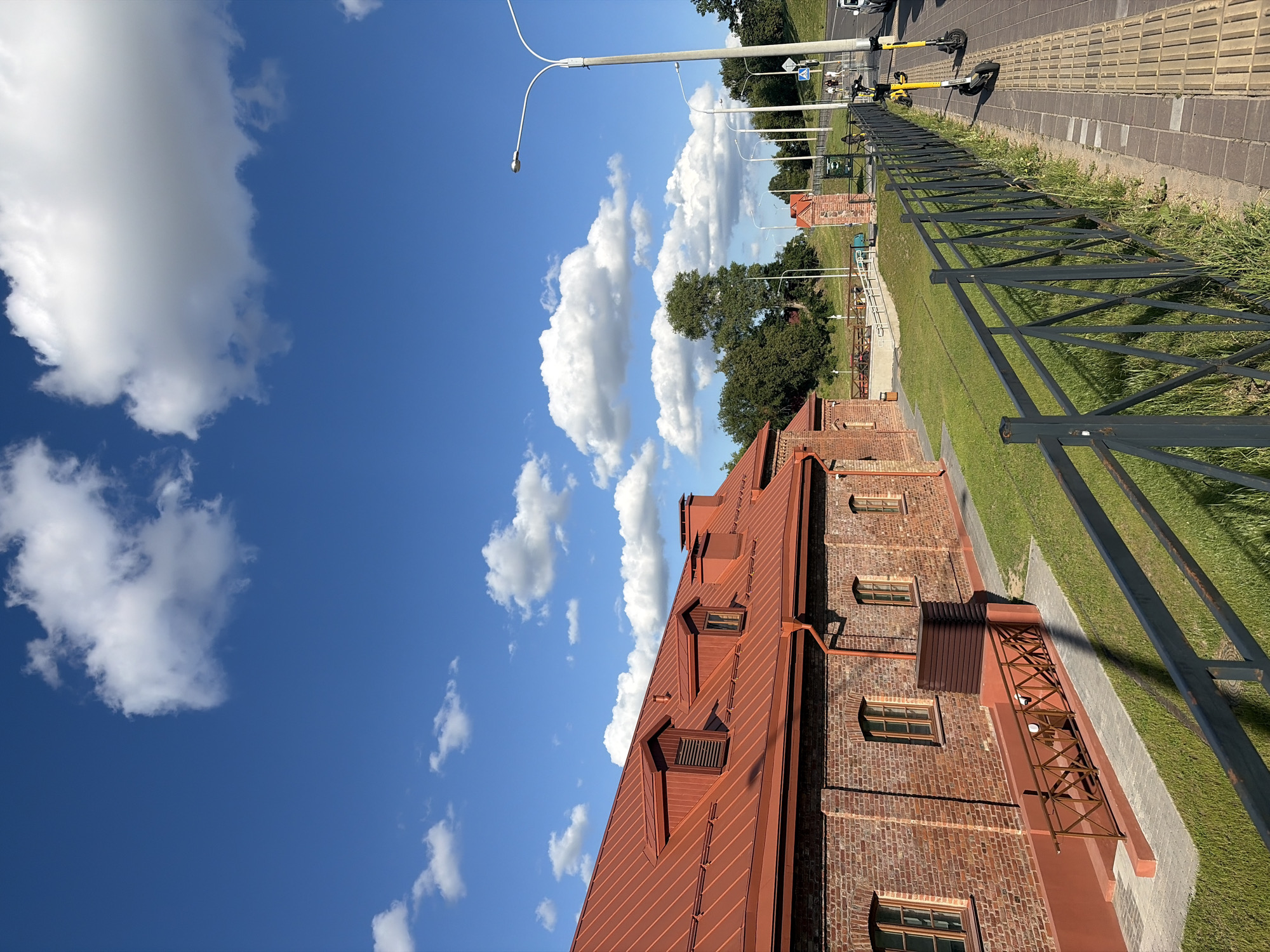 This media file depicts an outdoor scene in Minsk, Belarus, under a bright blue sky with scattered white clouds. The image is oriented vertically, with the sky occupying the left portion and the ground and buildings on the right.
The foreground features the corner of a red-brick building with a distinctive red metal roof. The roof has several dormer windows and a visible gutter system. Adjacent to the building is a grassy area bordered by a metal fence. Beyond the fence, a paved walkway is visible, with several yellow electric scooters parked on it. A street lamp with a curved arm extends into the frame from the top left. Further in the background, more trees and the tops of other buildings, including a smaller red-brick structure with a tower-like element, can be seen. A road with a few vehicles is also present in the distance.
The scene appears to be set during the daytime, given the bright sunlight casting shadows and the clear sky. The weather seems pleasant and sunny. There are no people actively engaged in activities, but the presence of parked scooters suggests a location that may be used for recreation or transportation. The overall mood is one of a calm, bright, and possibly urban setting. No visible text is present in the image.