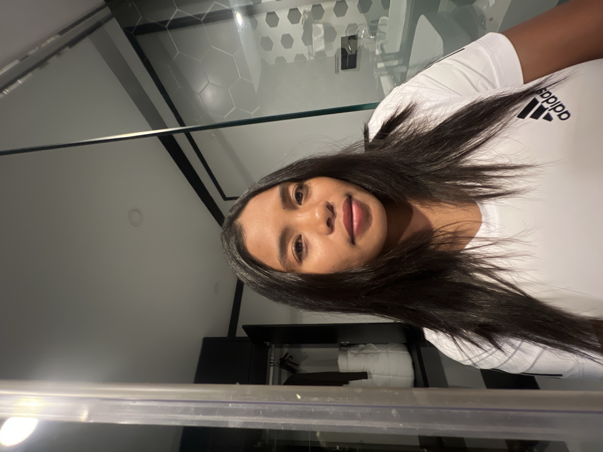 A young woman with long, dark hair is taking a selfie in what appears to be a hotel bathroom. She is wearing a white Adidas t-shirt. To her left, there is a clear glass partition, and beyond that, a black wardrobe with clothes hanging inside. To her right, a glass shower enclosure is visible, with hexagonal tiles on the wall and a sink and faucet in the background. The lighting is dim, with a bright light source in the upper left corner, possibly a bathroom fixture. The setting suggests a modern, clean hotel room.