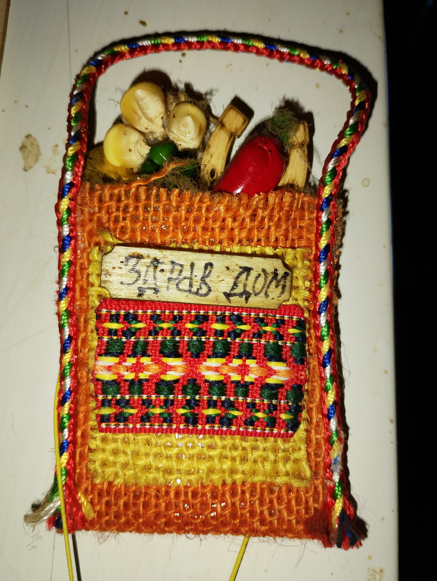 The image shows a handmade decorative item, likely a small pouch or basket, made from woven fibers and threads. It is filled with what appear to be miniature decorative items representing produce, such as corn kernels, a red chili pepper, and other small wooden or dried elements. A small wooden tag is attached to the front of the pouch with an inscription that reads "ЗДРАВ ДОМ" in Cyrillic script, which translates to "Healthy Home" or "Good Health to the Home." The item is adorned with a colorful braided handle made of red, blue, yellow, and green threads. The pouch itself is primarily orange and yellow, with a decorative woven band featuring red, green, yellow, and dark blue patterns. The item is placed on a light-colored, possibly white, surface that appears to be textured and has some faint brown stains or smudges. The lighting suggests it might be an indoor setting, but there are no identifiable features of a specific city or location.