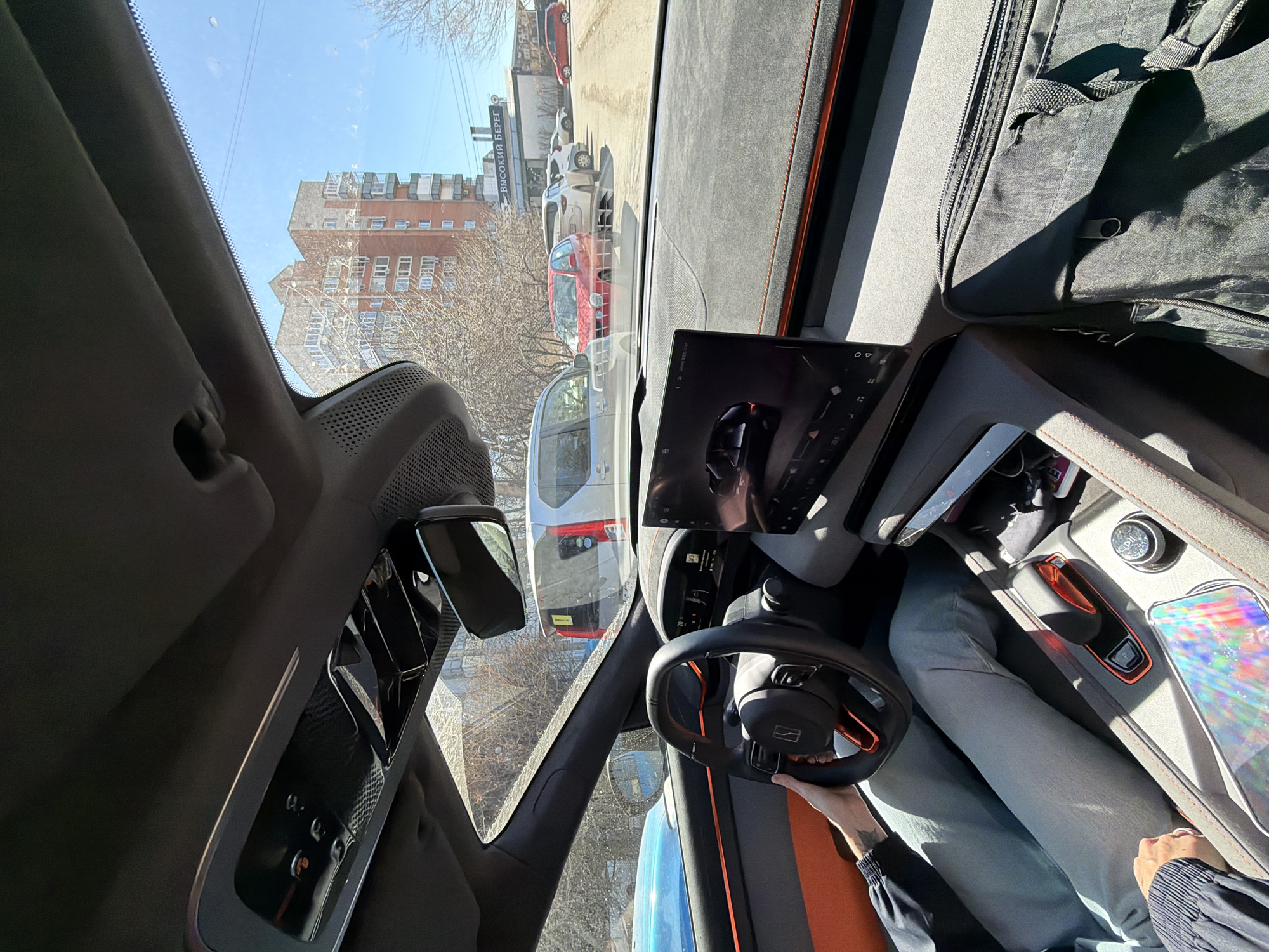 The image, captured from inside a modern car, presents an angled view of an urban street on a bright, sunny day. The perspective is from the driver's seat, looking outwards and slightly downwards.
Inside the vehicle, a person's hands grip a black steering wheel, which features orange accents and a distinct logo, identifiable as belonging to the Chinese EV manufacturer Li Auto (likely an L7 or L8 model). The dashboard is upholstered in a dark, possibly suede-like material with orange stitching. A prominent central infotainment screen displays a graphical interface, possibly a car rendering or navigation map. The center console, also adorned with orange accents, includes various controls such as a rotary dial, and what appears to be a mobile device reflecting holographic-like patterns. Another hand is visible resting on the center console. A rearview mirror and part of a side mirror are visible, as is a black bag or piece of luggage on the passenger side.
Outside the car, the street is lined with parked vehicles, including a red hatchback, a white van, and a grey SUV. In the background, a multi-story brick building with numerous windows stands under a clear blue sky. Bare tree branches suggest late winter or early spring. A white sign visible on a building across the street displays Cyrillic text: "Высокий Берег" (Vysoky Bereg), which translates to "High Bank" or "Tall Shore." This text confirms a Russian-speaking location, consistent with the provided context of Krasnoyarsk, Russia. The scene depicts a modern car stationary on a sunny city street.