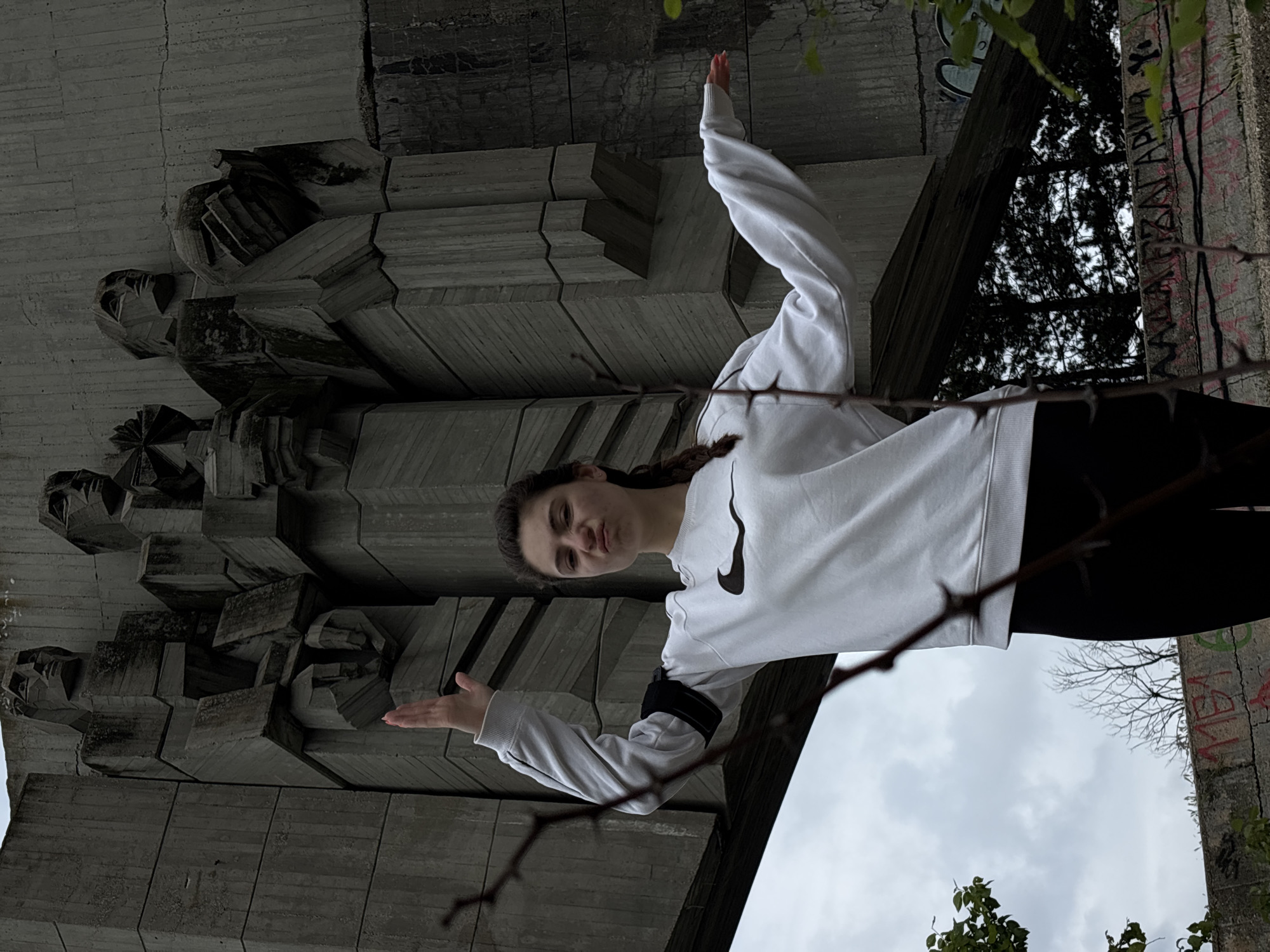 The image depicts a woman standing beneath the Monument to the Soviet-Bulgarian Friendship in Varna, Bulgaria. She wears a white Nike sweatshirt and black leggings. The massive concrete structure of the monument, adorned with stylized depictions of faces, looms overhead. The sky is overcast. Branches with thorns appear in the foreground. Graffiti is visible on the side of the monument. The photograph is taken from a low angle, looking up. The image is rotated approximately 90 degrees clockwise.