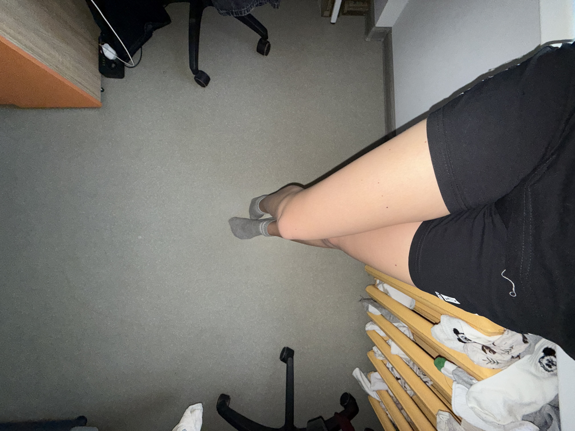The scene is a top-down view of a room. The floor is gray. A person wearing black shorts and gray socks is present. A desk with an orange edge and two office chairs are visible. A clothes drying rack with clothes hanging on it is also present. The location is not discernible from the image, and there is no indication that it is in Perm, Russia.