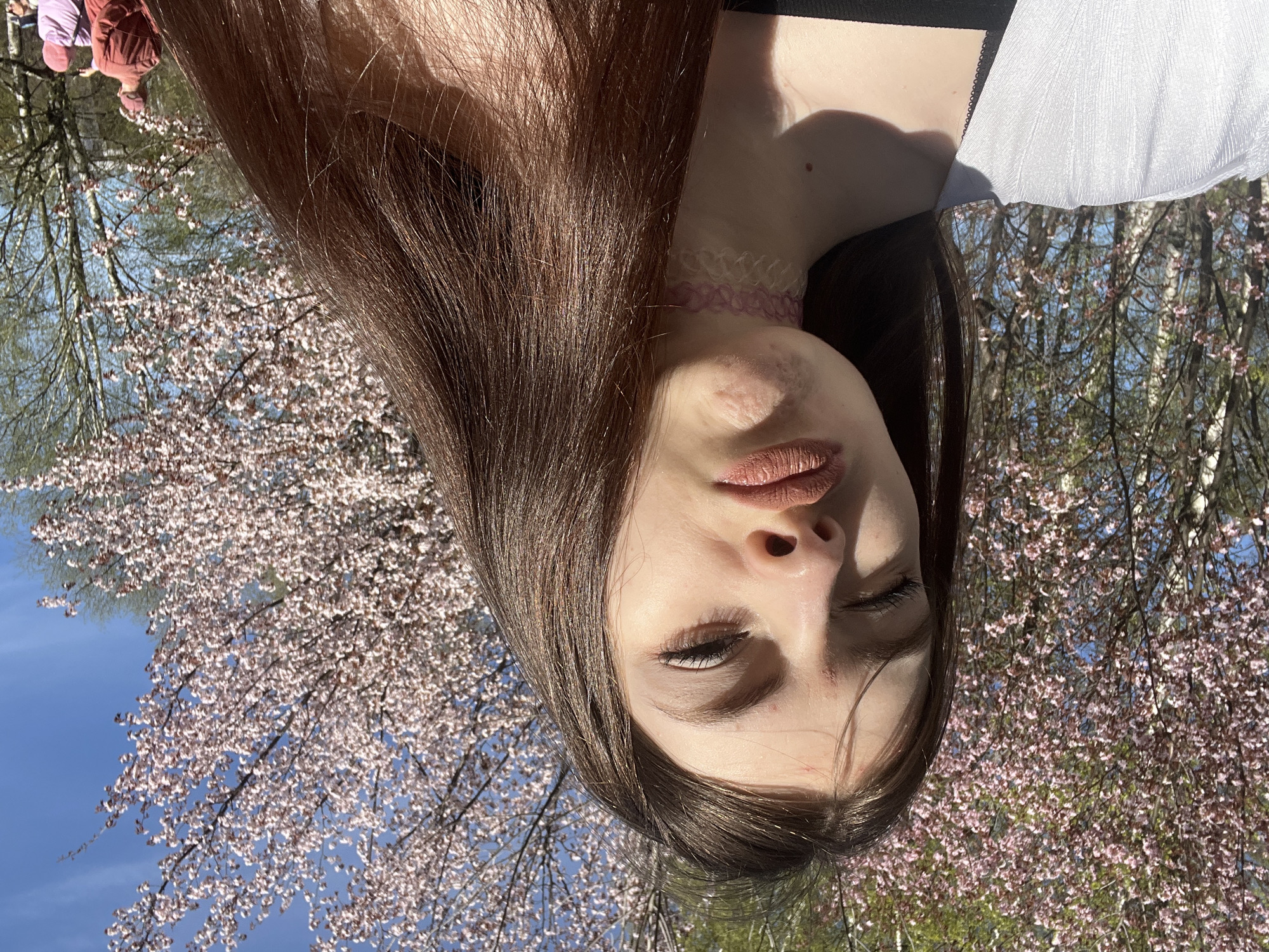A young woman with long brown hair is posing for a selfie in front of blossoming trees in an area inferred to be in Unknown, Russia. She is wearing a pink choker. The background shows flowering trees against a blue sky. Two people are partially visible in the upper left corner of the image.