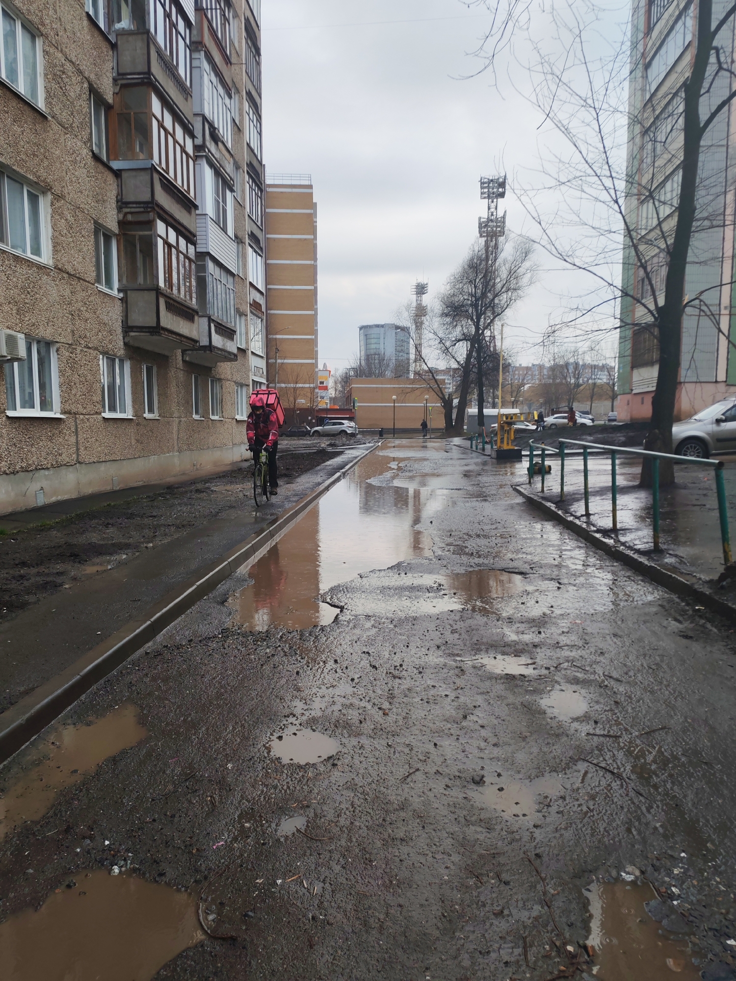 The image shows a street in Izhevsk, Russia, heavily damaged with potholes filled with muddy water. On the left, a multi-story apartment building with weathered beige concrete panels is visible. A cyclist wearing a red jacket and a large pink delivery backpack is riding along the edge of the road, navigating the potholes. In the background, additional buildings are visible, including a tall tan-colored building and communication towers. Bare trees suggest it is late autumn or early spring. A green metal railing runs along the right side of the flooded street. The sky is overcast and gray. Several vehicles and pedestrians are visible in the distance.