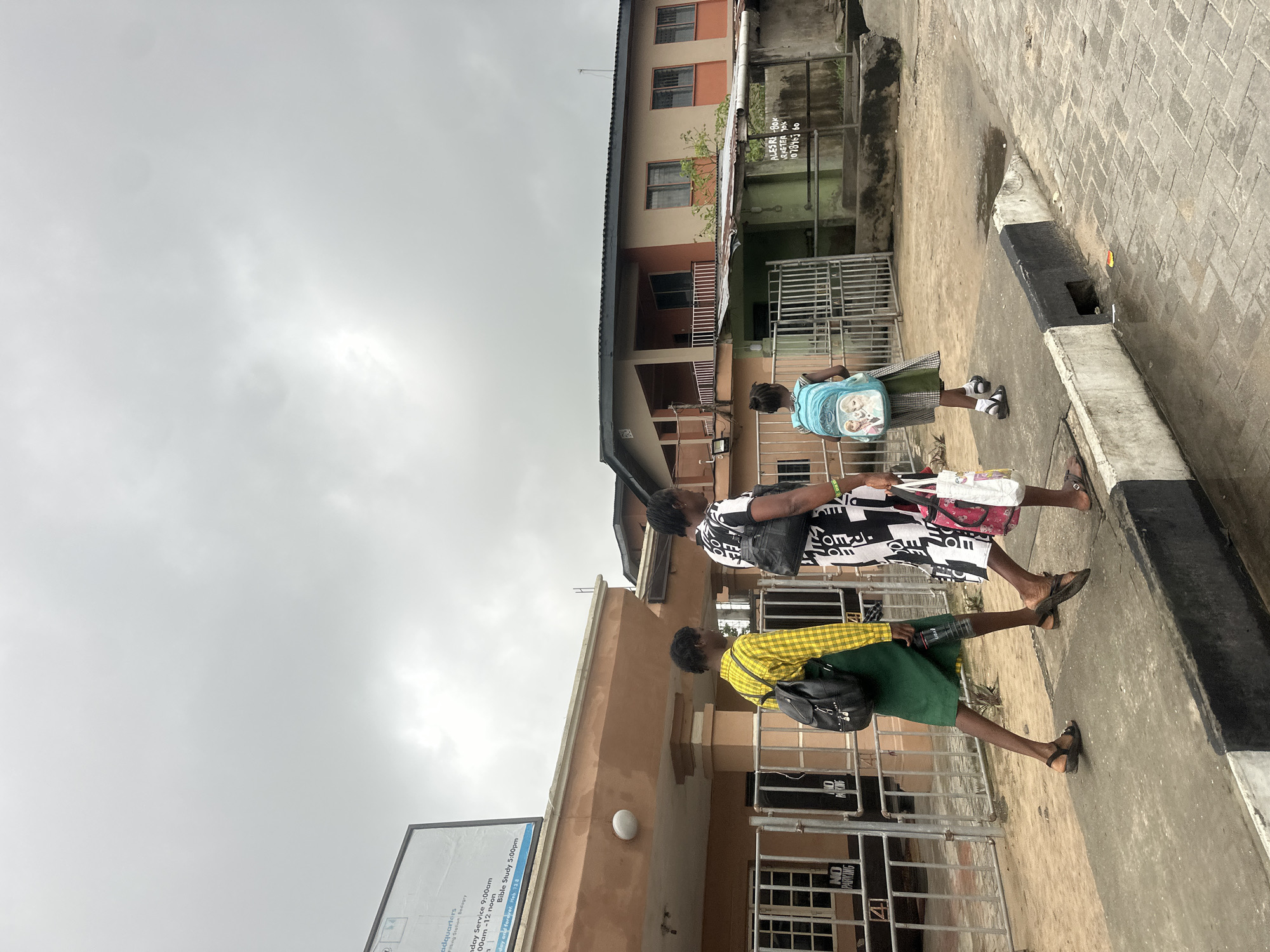In Badagry, Nigeria, under a cloudy sky, three individuals walk along a paved sidewalk. The person in the foreground, on the left, wears a yellow plaid long-sleeved shirt and a green skirt, carrying a black backpack and a water bottle. To their right, a person in a black and white patterned dress walks, holding a black handbag and a pink and white shopping bag. On the right side of the group, a child wearing a light blue backpack with cartoon characters on it, a green and white plaid skirt, and white socks walks ahead of the others. They are passing a fenced area in front of a light brown building with windows and a sign at the top left. Beyond the initial building, another building with orange and beige sections is visible, with a green, shed-like structure in front of it. The environment appears to be an urban street with scattered puddles on the ground, suggesting recent rain.