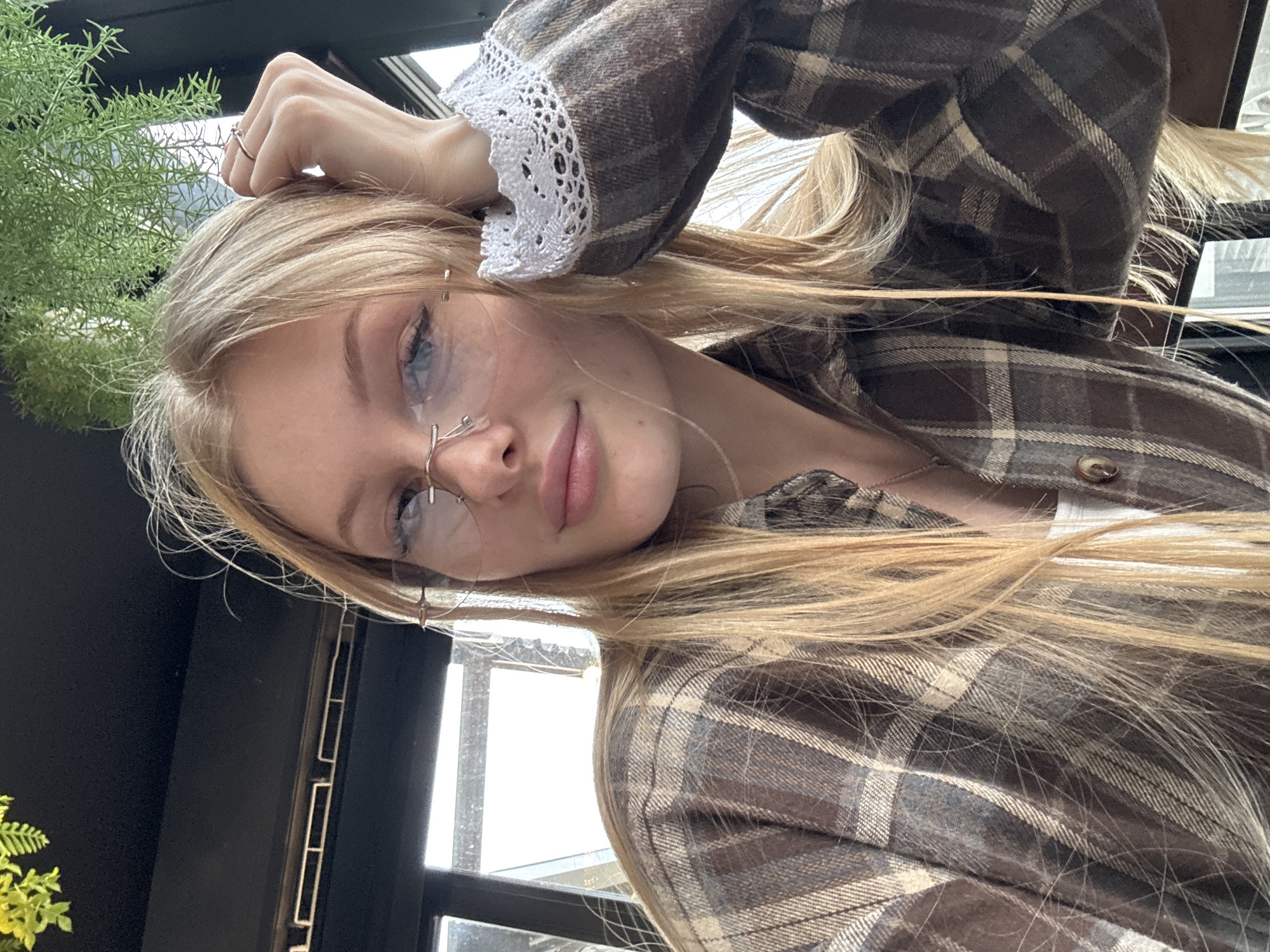 A young woman with blonde hair and fair skin poses for a close-up selfie indoors. She is wearing wire-rimmed glasses and a brown plaid shirt with white lace trim on the sleeve cuffs. Her hand rests on her head, displaying a ring. To the left, a window frame and greenery are visible. The image is taken at an angle, making it difficult to pinpoint the exact location in Perm, Russia, but the setting appears to be a cafe or restaurant with a view.