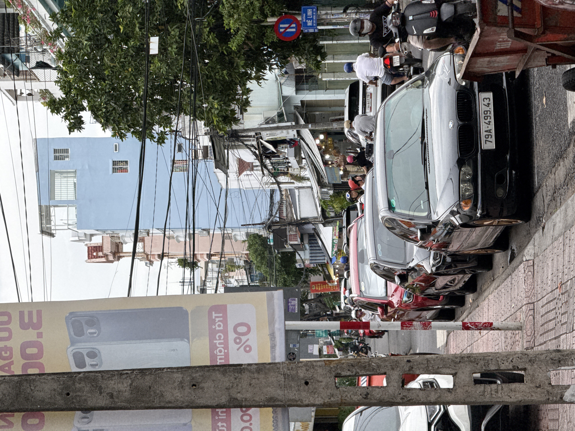 This is a street scene in Nha Trang, Vietnam, featuring a traffic jam with cars and motorcycles. On the left, a large banner displays an advertisement, and a concrete utility pole stands in the foreground. Several cars are lined up on the right side of the road, including a black BMW with the license plate "79A-499.43". Behind the BMW, a scooter is visible. People on motorcycles are navigating through the traffic. The buildings in the background are typical of an urban environment, with power lines crisscrossing overhead.