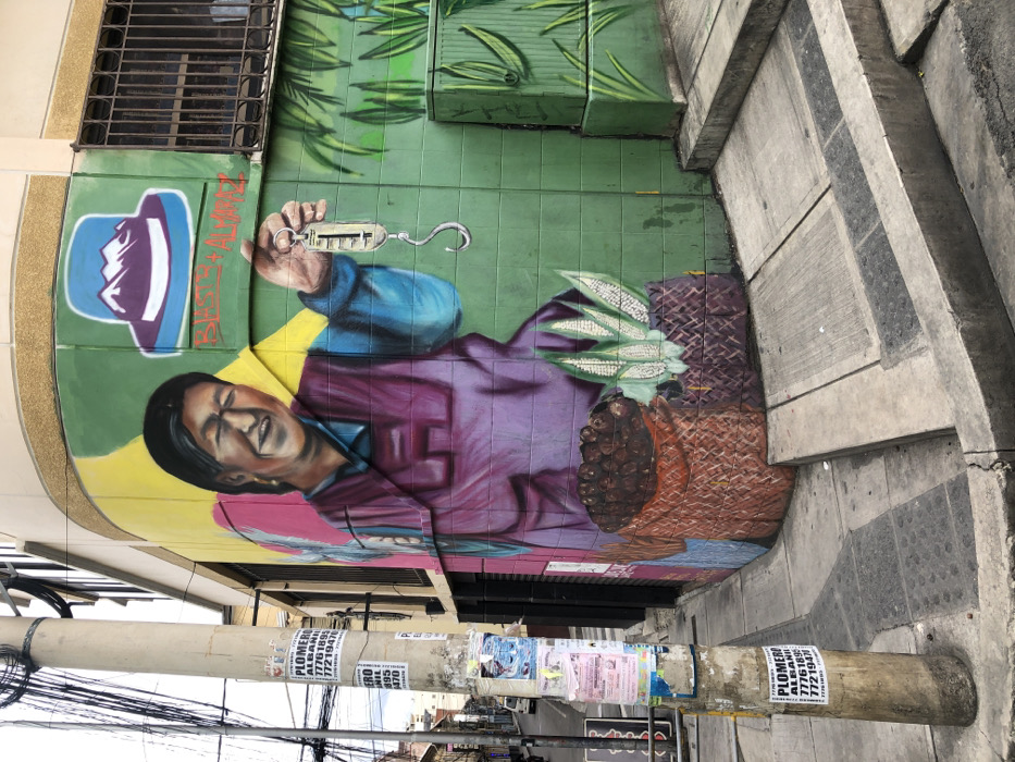 A street mural depicts a smiling woman in a purple shirt and blue sleeves, holding a scale with a hook. Next to her is a woven basket containing corn and coffee beans. Above her, a blue hat with mountains is painted. The mural is on a green tile wall next to concrete steps. A metal grating is visible at the top left. Utility poles covered with flyers are in the foreground, leading to a distant street scene.