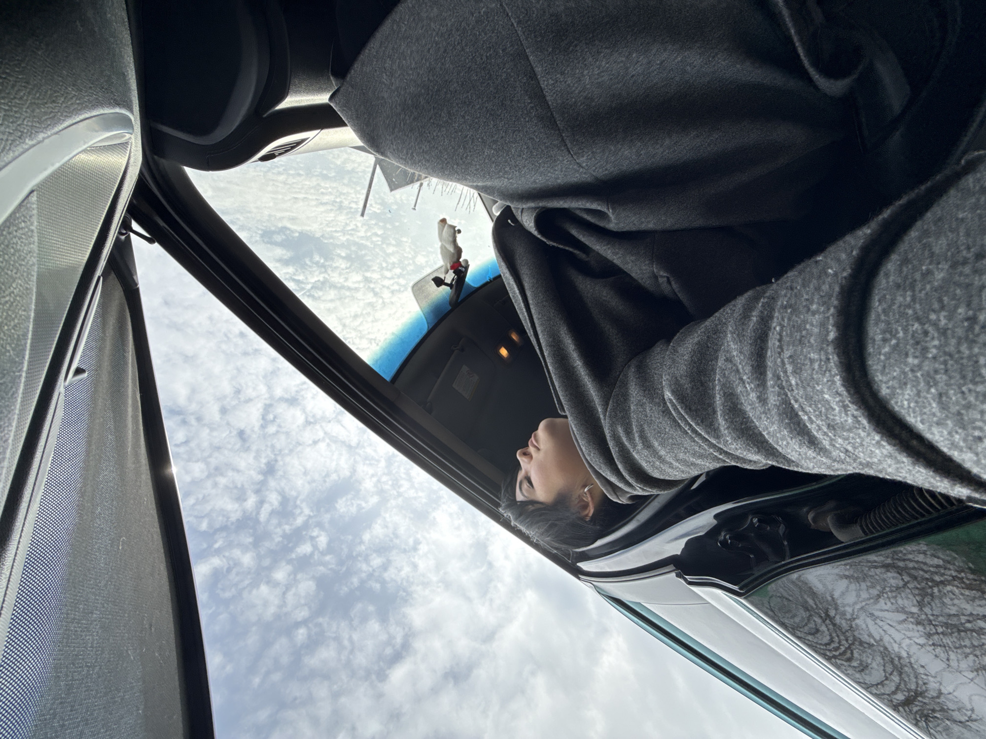 A low-angle view looking up through the open sunroof of a vehicle. A person wearing a gray hooded garment is partially visible, leaning into the sunroof opening. The sky is visible through the sunroof, showing white clouds. A small doll is attached to the windshield. The interior of the car is visible, including the sunroof controls and the vehicle's interior trim. It's impossible to verify the location in the city of Chaikovsky, Russia, without additional information or context.