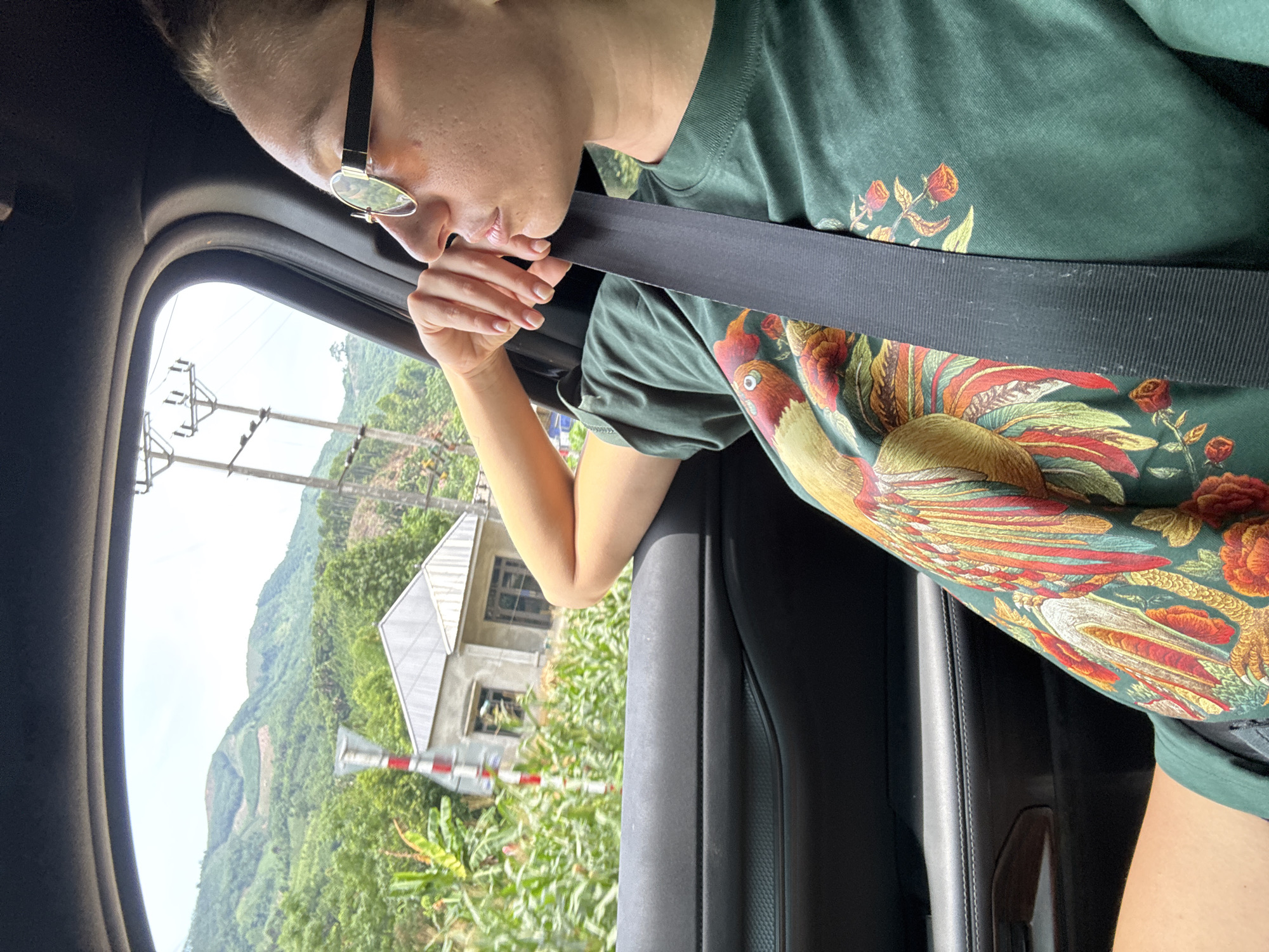 An eye-level shot from inside a vehicle captures a person looking out the window at a rural landscape, likely in Bảo Hà, Vietnam. The main subject, appearing to be female, is wearing dark-rimmed glasses and a dark green t-shirt adorned with a vibrant, colorful rooster or bird and floral pattern. She is buckled into a seatbelt and is holding the strap with her right hand, leaning forward to look outside. Her expression is partially obscured, but she appears to be observing the view intently.
Through the car window, a lush, mountainous terrain covered in dense green foliage is visible under a bright, somewhat hazy sky. Closer to the foreground, a simple, unfinished-looking house with a grey roof and unrendered concrete walls stands amidst greenery. Power lines and utility poles are visible against the sky, and a red and white striped railroad crossing barrier is prominent near the house, indicating proximity to train tracks. The immediate car interior shows part of a dark car door and window frame. The overall scene suggests a journey through a scenic, possibly remote, area during daylight hours. No visible text is present in the image.