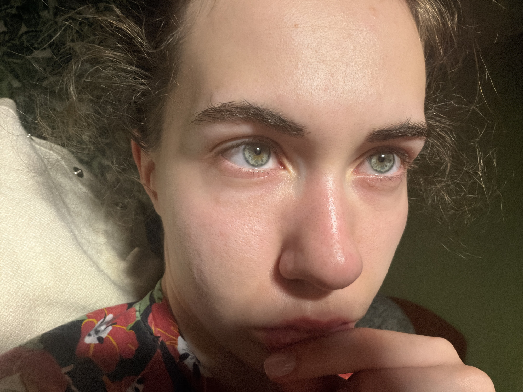 A close-up, head-and-shoulders shot shows a person with light skin and light brown, wavy hair. The person's green eyes are looking to the right. Their eyebrows are dark and well-defined. The skin on the face appears slightly red, particularly on the cheeks and around the nose. The person's right hand is resting on their mouth, with their thumb touching their lower lip. They are wearing a dark shirt with a floral pattern featuring red flowers and green leaves. The background is dark, with some lighter areas that suggest an indoor environment with soft lighting. There is no visible indication of the city of Zavodoukovsk, Russia.