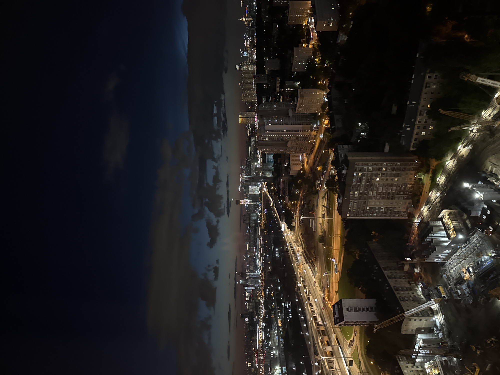 The image captures an aerial view of a cityscape at night, with a dramatic sky transitioning from deep blue to twilight hues on the left. The right side of the image showcases a densely populated urban area with numerous illuminated buildings of varying heights. A major highway or elevated road network is visible, with car headlights creating streaks of light. Construction sites with cranes are also present, indicating ongoing development. Trees and green spaces are interspersed within the built environment.
The scene depicts a bustling city at night, likely during a period of clear weather with some cloud cover. The time of day is clearly night, as evidenced by the artificial lighting illuminating the city and the dark sky. The presence of a highway and numerous residential and commercial buildings suggests a large urban center. The construction activity hints at a growing or developing area.
While no people are discernible, the numerous lights suggest a significant population. There is no discernible text in the image. The overall impression is one of a dynamic and active urban environment at night, with elements of both established infrastructure and ongoing construction.