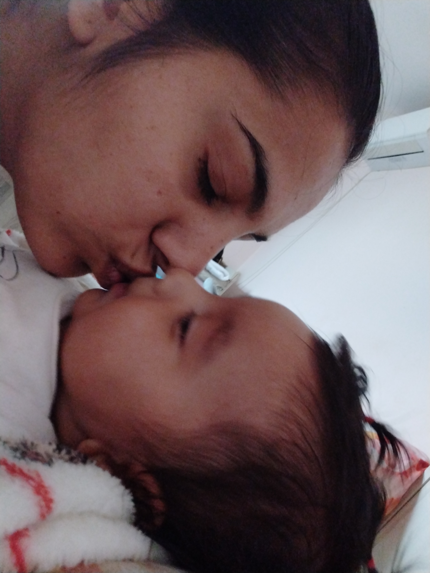 A close-up shot depicts a woman kissing a baby. The woman has dark hair pulled back and closed eyes. The baby lies on a blanket and has dark hair. The location is Unknown, Unknown.