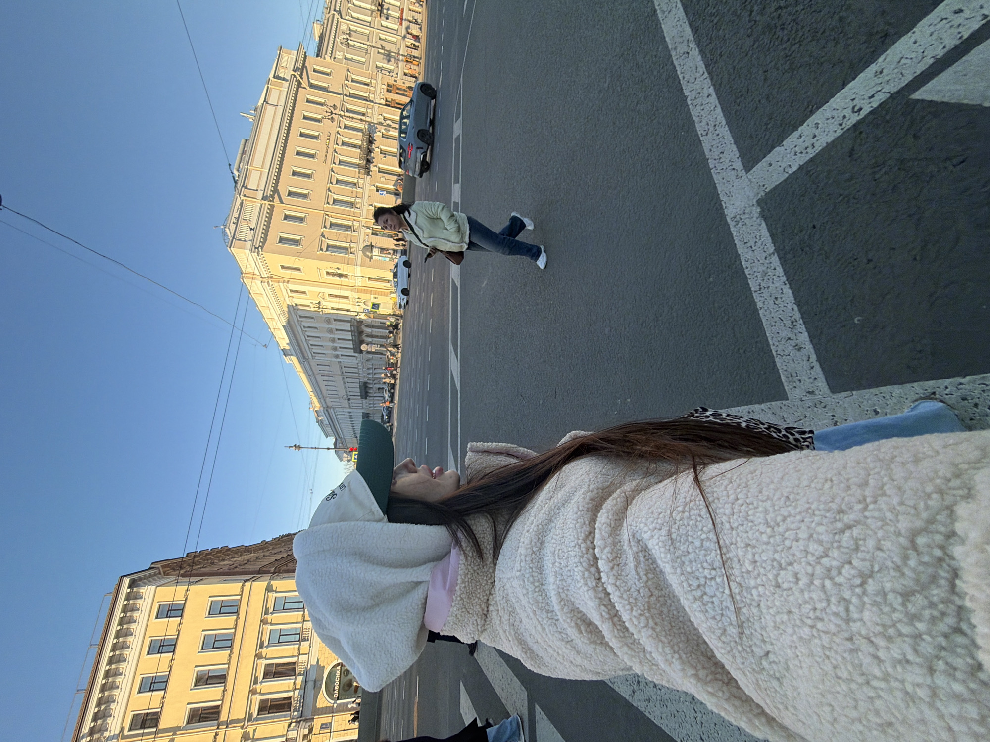 The image captures a sunny street scene in Saint Petersburg, Russia. A woman wearing a white sherpa jacket and a green baseball cap is lying down on a pedestrian crossing with prominent white markings. In the background, another woman is seen walking across the street. Cars are parked and moving on the street. The buildings flanking the street are tall, ornate structures typical of the city's architecture. The sky is a clear blue.