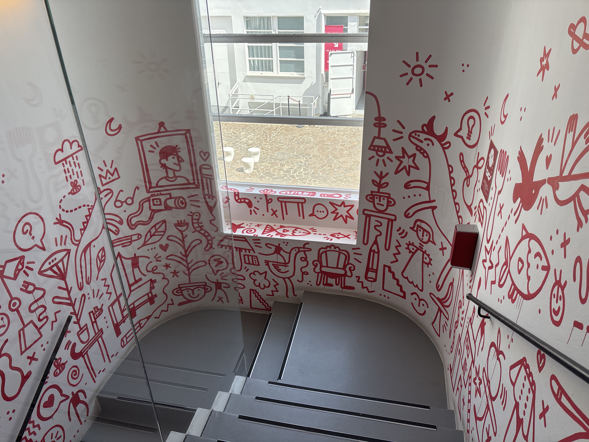 This media file shows a white staircase in Milan, Italy, decorated with whimsical red doodles. The staircase has gray treads and risers, with a black handrail on the right. A glass railing runs along the left side of the stairs. The walls are covered with a variety of cartoonish red drawings, including a portrait in a frame, a smiling fish, a bird, a dinosaur-like creature, a lamp, a plant in a pot, a chair, and various abstract shapes and symbols like clouds, hearts, and stars.
A large window is visible at the top of the staircase, looking out onto an outdoor courtyard paved with irregular stones. Through the window, a white building with multiple windows and a red sign is visible. Three white cylindrical stools are also present in the courtyard. The sunlight suggests it is daytime, and the lighting indicates pleasant weather. The overall impression is one of a bright, artistic, and playful space. There is a small red fire alarm box mounted on the wall to the right of the window. The red sign visible through the window reads "SALA DI RICEVIMENTO".