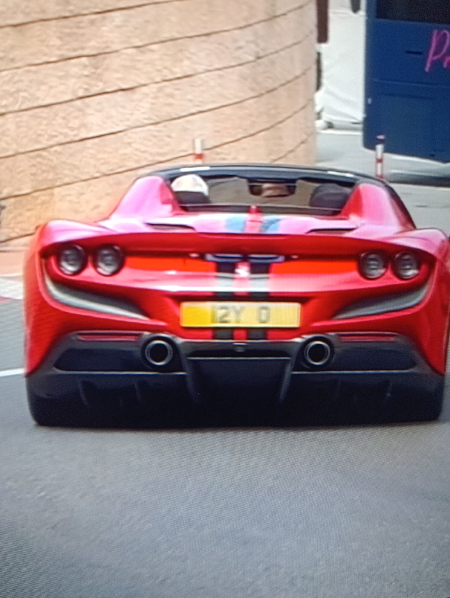 The image shows the rear of a red Ferrari F8 Spider with a yellow license plate reading "12Y 0." The car has a black and gray diffuser, round taillights, and dual exhaust pipes. A blue and white stripe runs along the center of the car's rear. The background includes a tan building and a blue bus with the letters "Pr" visible. The location appears to be a city street.