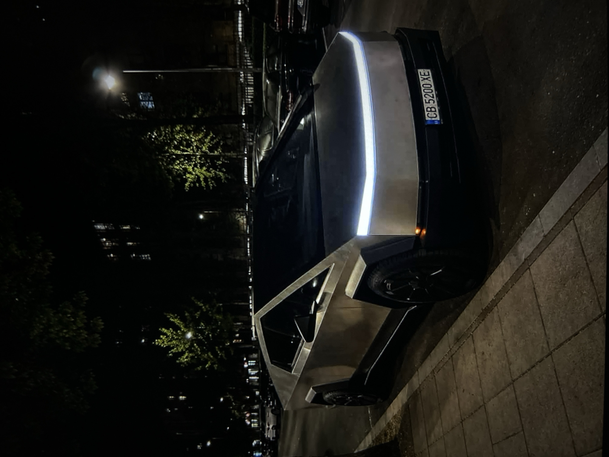 A silver Tesla Cybertruck is parked on the side of a street at night. The vehicle is illuminated by streetlights and its own headlights, which are a bright white strip across the front. The car has a distinctive angular, futuristic design with sharp edges and flat planes. The license plate reads "CB 5200 XE." The street is paved with cobblestones on the sidewalk and asphalt on the road. In the background, there are trees with illuminated foliage and buildings with glowing windows, suggesting an urban environment. Other cars are parked in the background, some of which are also illuminated. The overall scene is dark, with the primary light sources being artificial.