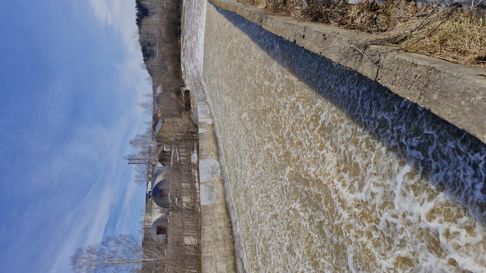 A landscape-oriented photograph displays a water cascade from an elevated concrete structure, resembling a dam. The photograph is rotated to the left.
The water, appearing frothy and brown, is flowing from top-right to bottom-left. The edges of the waterway are lined with weathered concrete structures. On the right, dry vegetation clings to the edge. On the left side of the water source are building structures that imply urban life.
Bare trees and buildings are visible in the background, on the left. Above them are streaks of white clouds against a blue sky. The lighting suggests a sunny day. The geographic location is identified as Otchor, Russia.